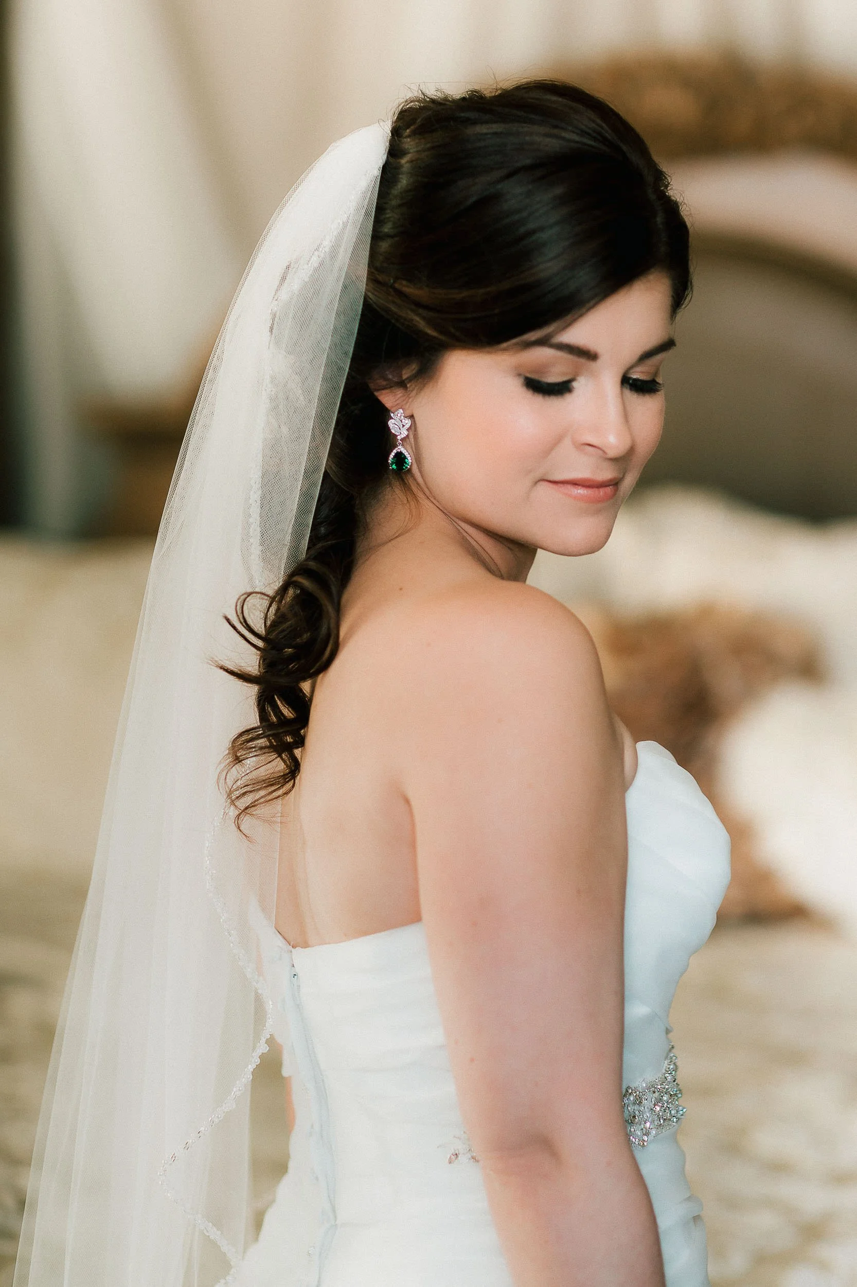 A bride with dark hair styled in loose waves, wearing a white strapless wedding gown with a bejeweled belt, and a long veil, looking down with a serene expression, in a softly lit indoor setting.