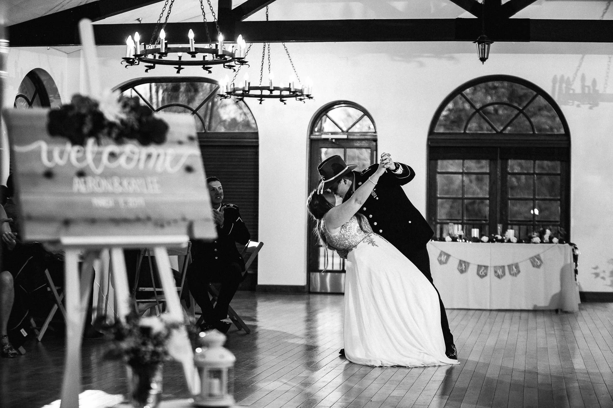 A bride and groom sharing their first dance at a wedding reception in a decorated hall with large windows and chandeliers.