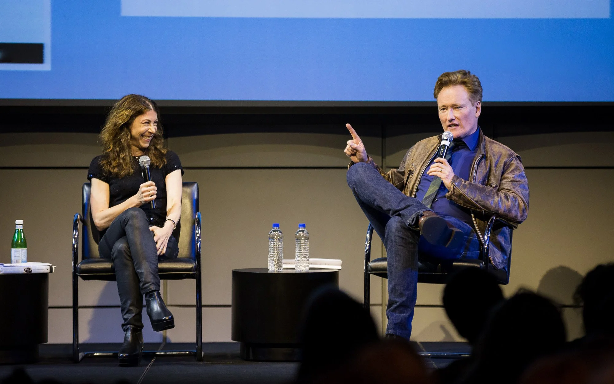 Two people seated on stage, a woman on the left and a man on the right, holding microphones and engaged in conversation. The woman is smiling, wearing black clothing and boots, and the man has reddish hair, wearing a leather jacket and jeans. They are in a panel discussion setting with water bottles on a small table in front of them.