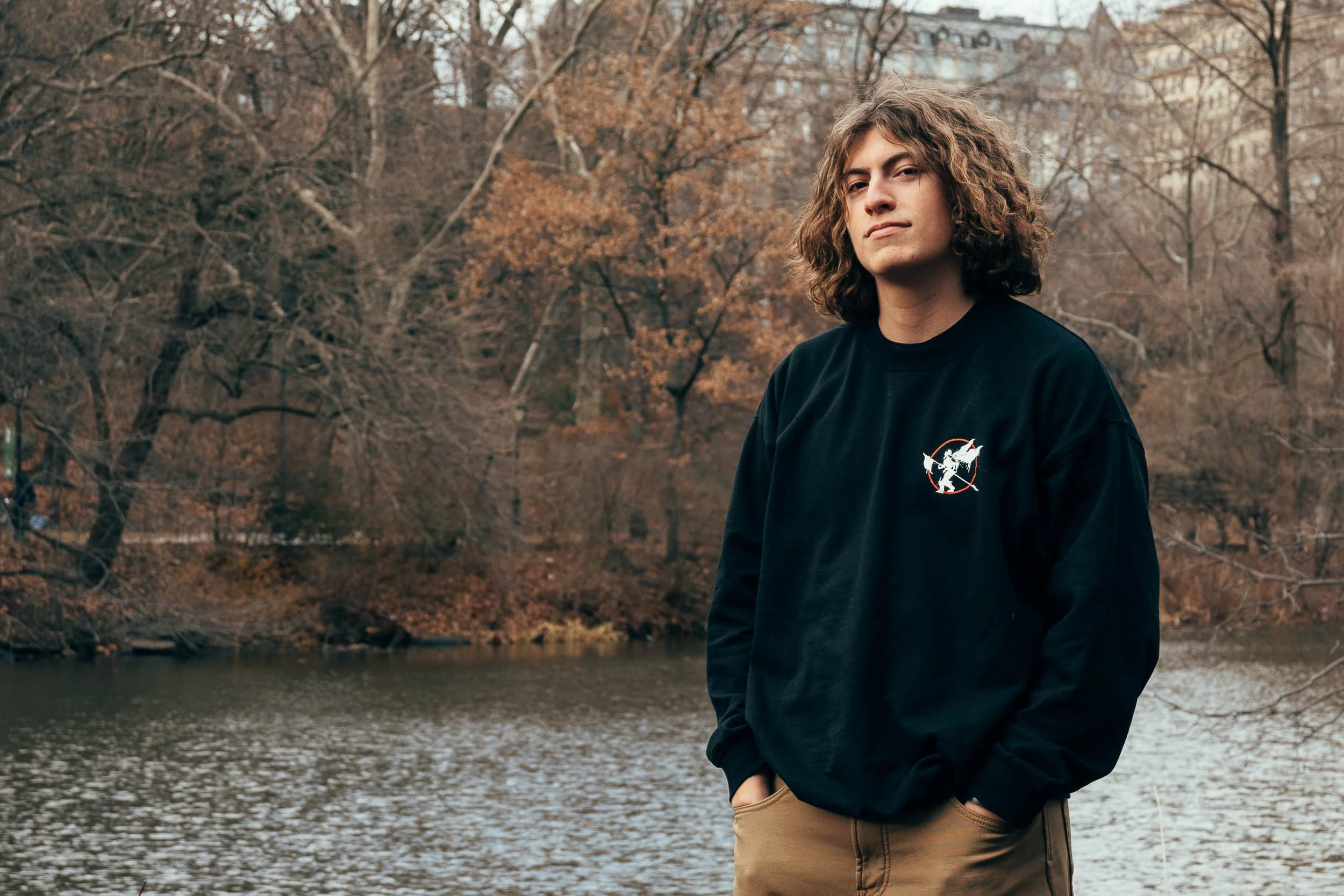 A young man with curly hair and a serious expression standing by a river in a wooded park during fall in New York, wearing a black sweatshirt with a graphic on it and beige pants.