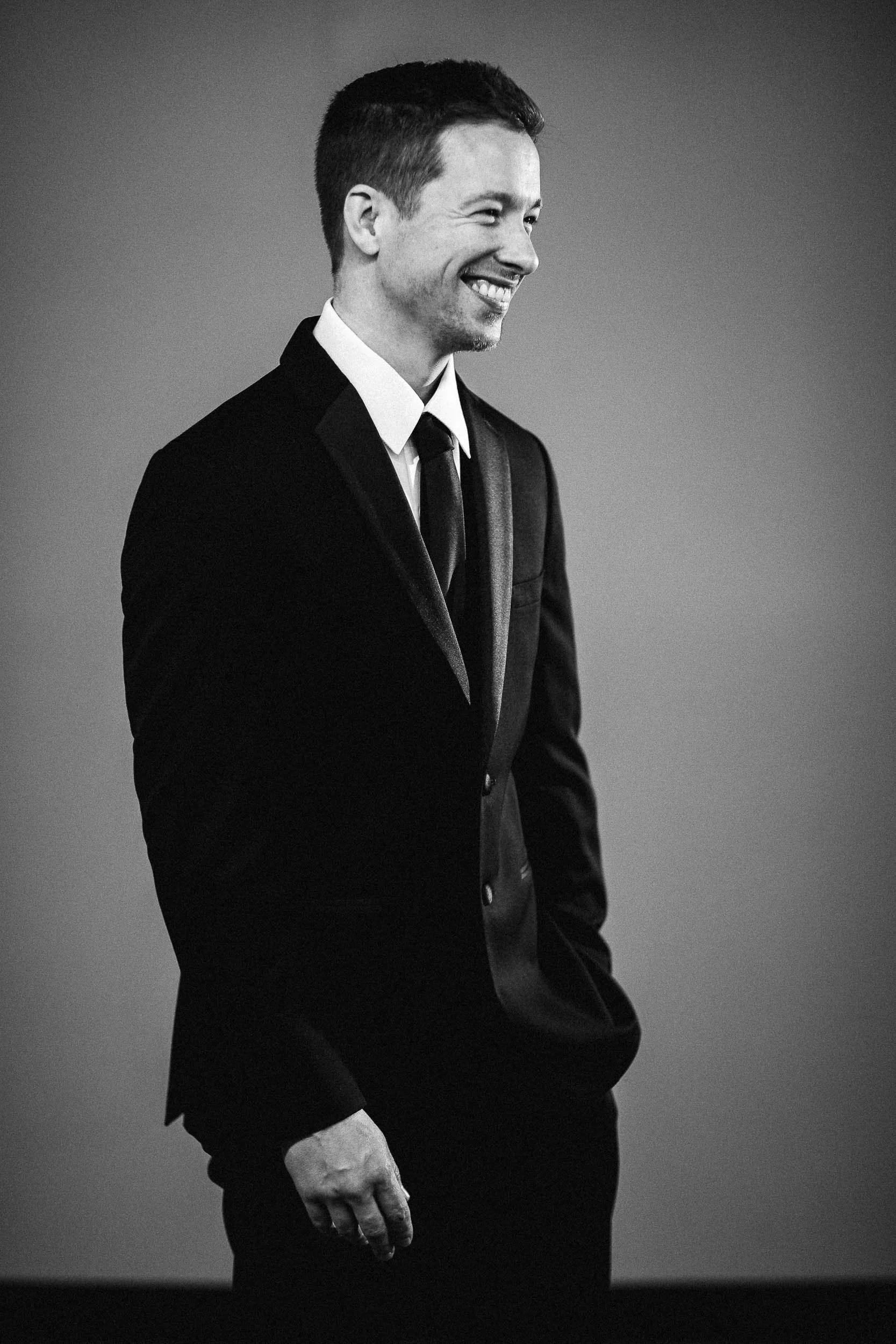 A black-and-white photo of a smiling man in a business suit against a plain background. He is shown from the chest up, with his left hand relaxed by his side.