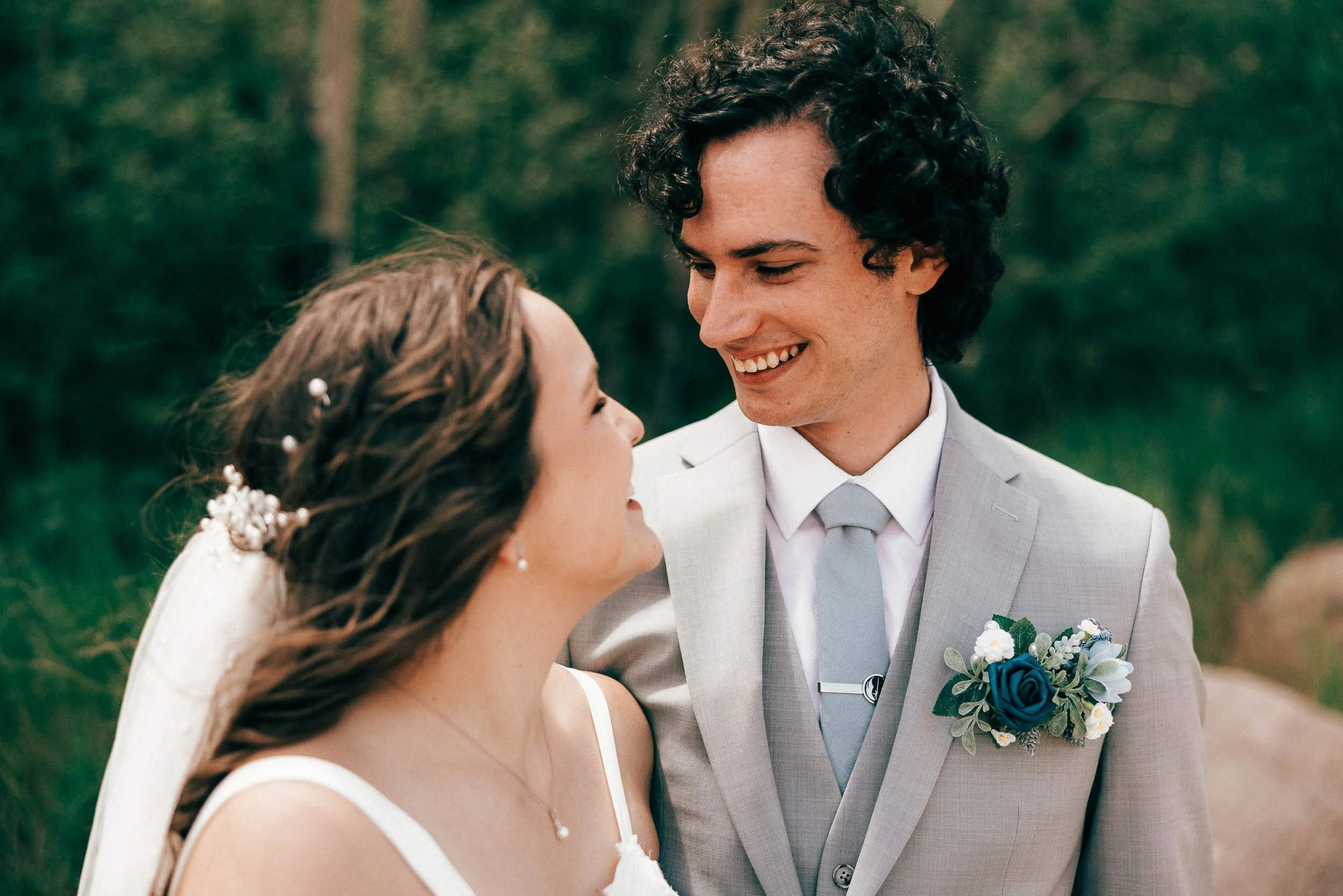 A bride and groom sharing a close moment outdoors, smiling at each other, with the bride wearing a white dress and floral accessories, and the groom in a light gray suit with a matching boutonniere and tie.