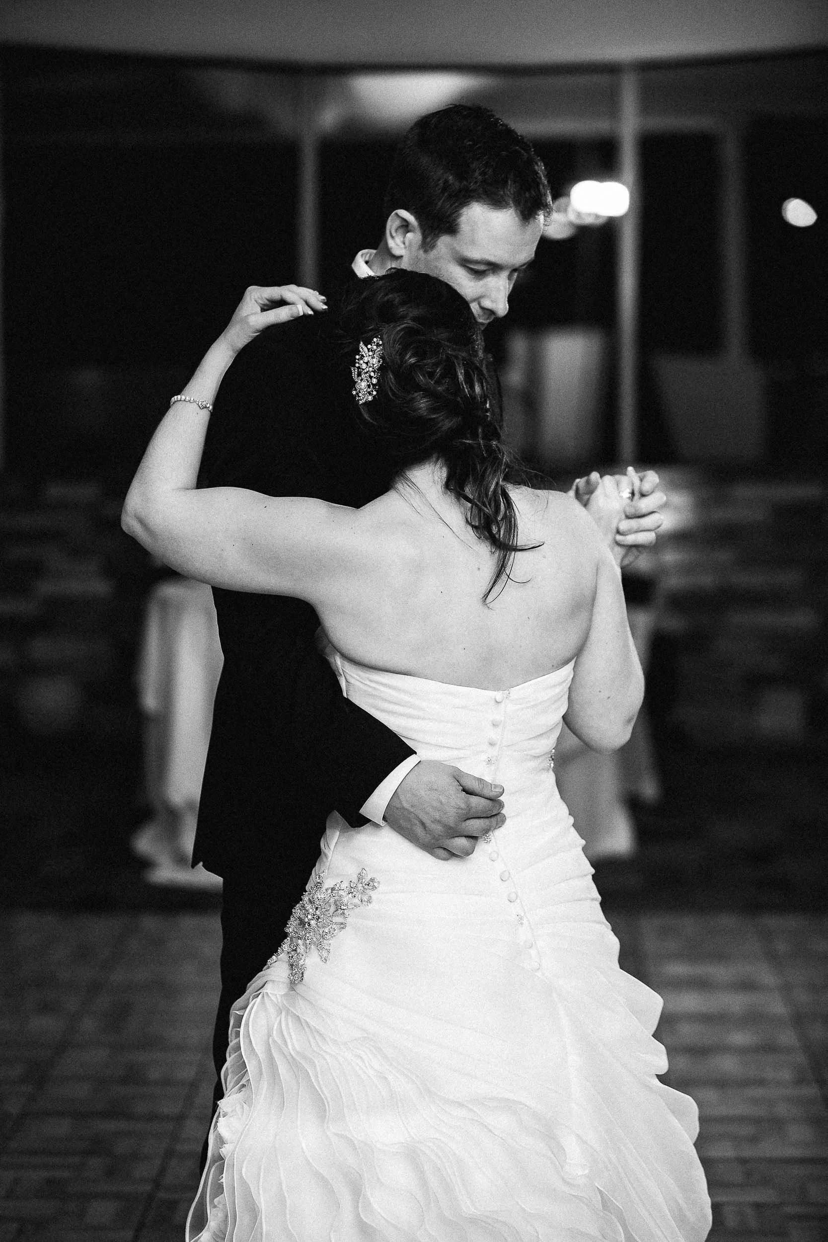 A black and white photograph of a couple, likely newlyweds, dancing closely together. The woman is wearing a strapless wedding dress with embellishments on the side and a decorative hairpiece. The man is dressed in a suit. They are holding each other