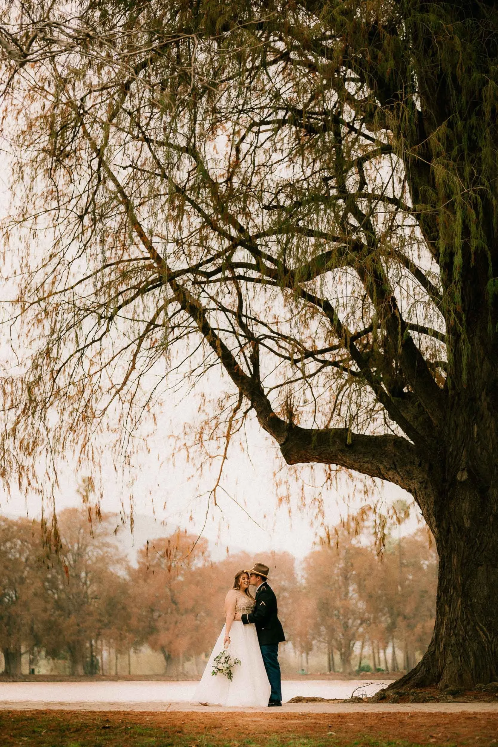 A couple dressed in wedding attire stands close together under a large tree in an outdoor park, surrounded by falling autumn leaves and blurred trees in the background.