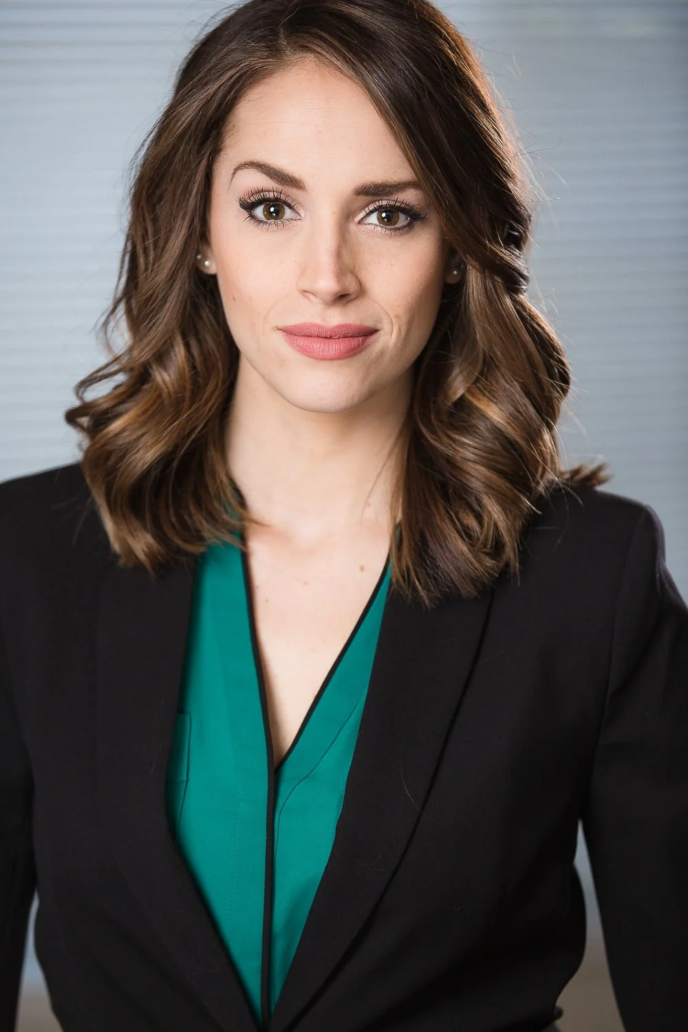 A woman with shoulder-length wavy brown hair, wearing a black blazer over a teal blouse, looking directly at the camera with a slight smile, in an indoor setting.