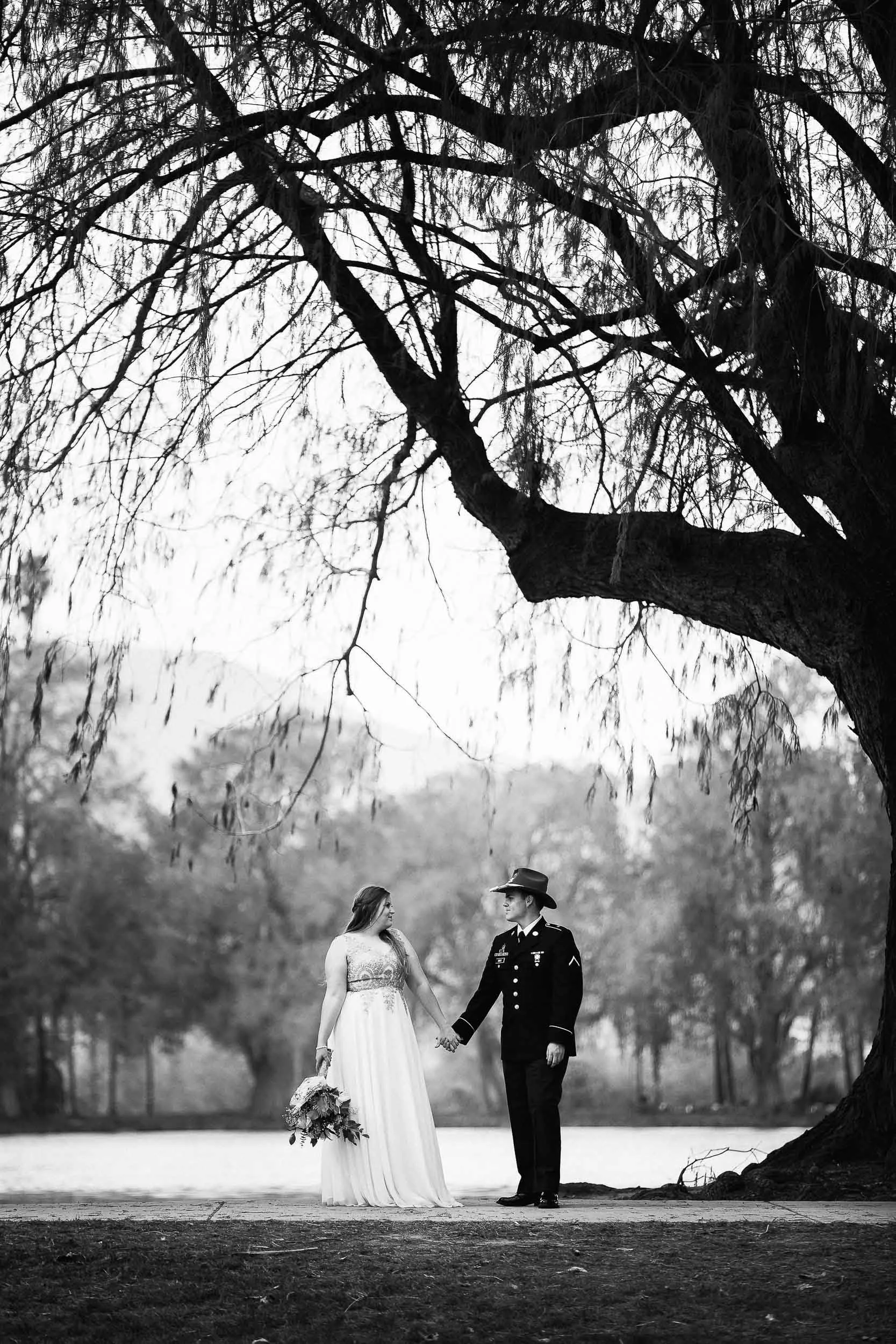 A black and white photo of a bride and groom holding hands outdoors, standing on a paved surface near a large tree with bare branches, with a lake and trees in the background.