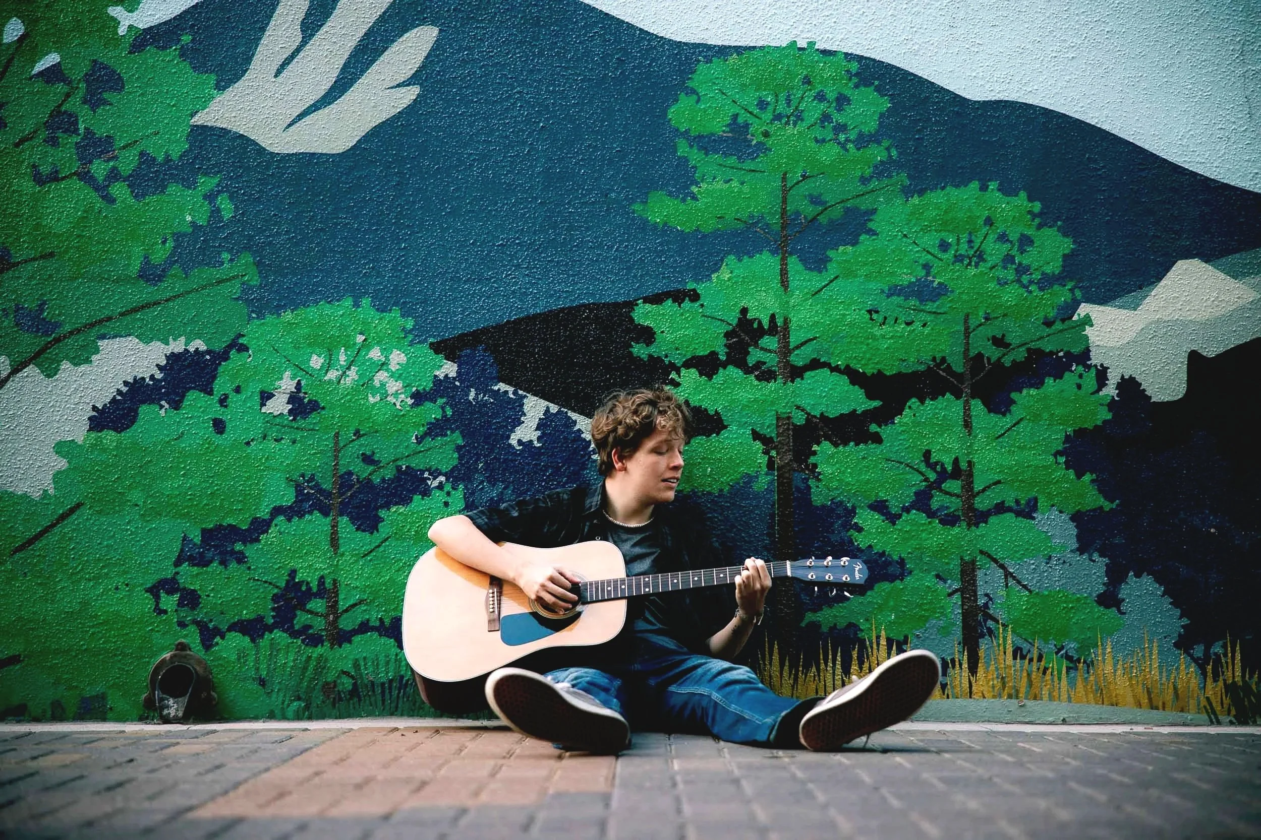 A young man with curly hair sits on the ground playing an acoustic guitar in front of a colorful mural of green trees and mountains.