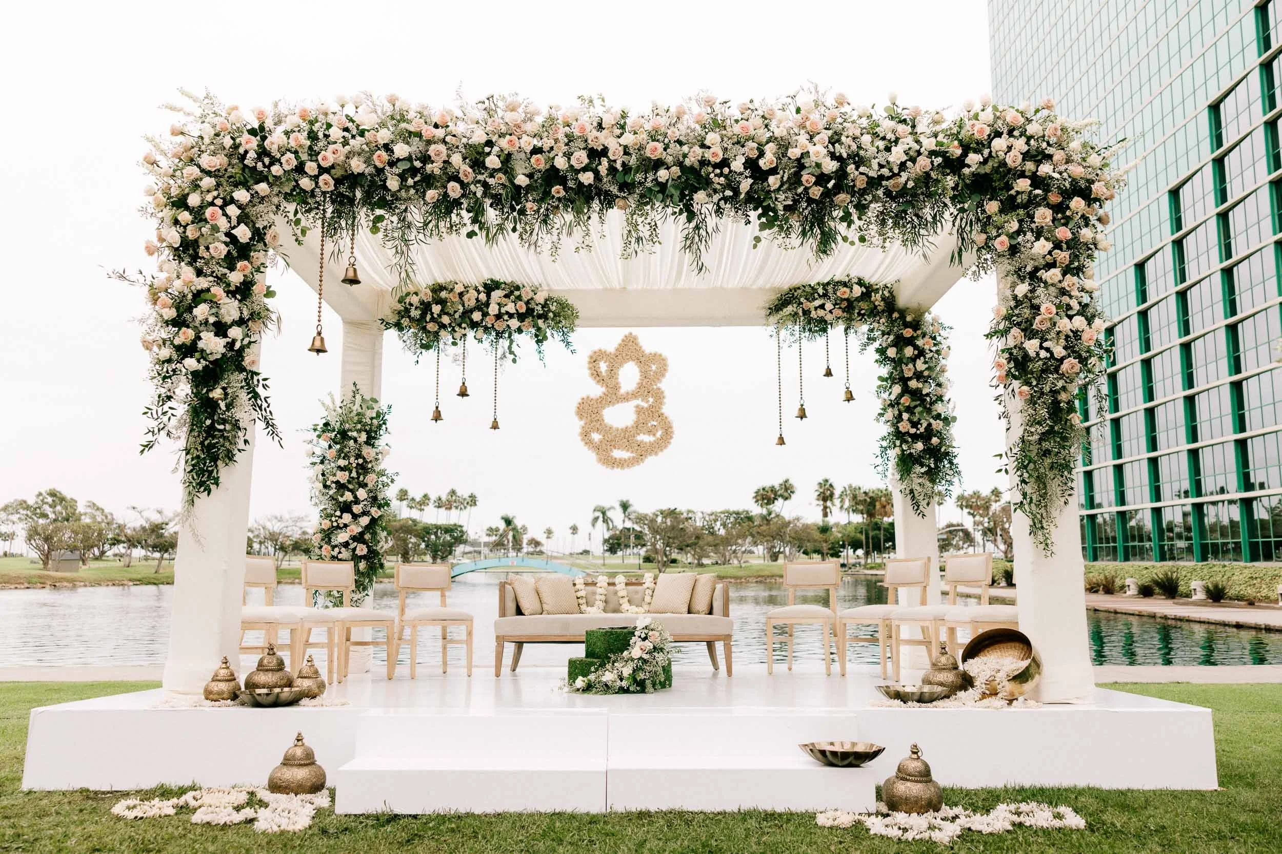 Wedding altar decorated with white and blush pink flowers, greenery, hanging bells, and a floral emblem backdrop, set outdoors near water.