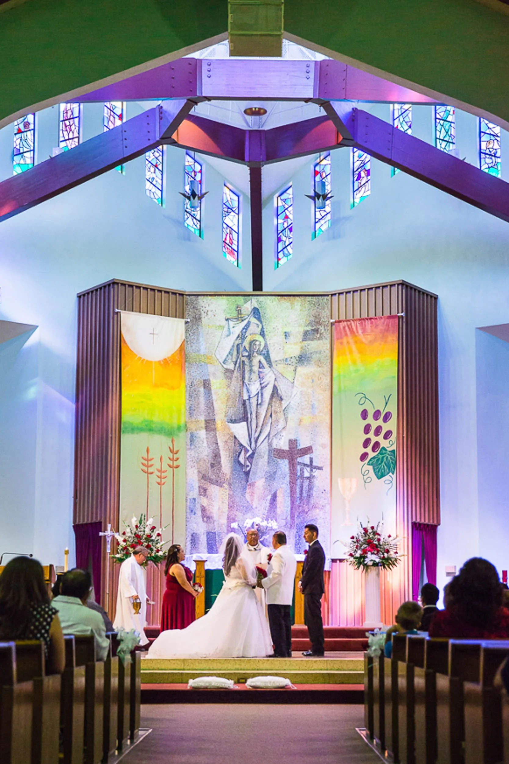 Inside a church during a wedding ceremony, with a bride and groom at the altar, surrounded by officiants and wedding guests. The church has stained glass windows, colorful banners, and floral arrangements.