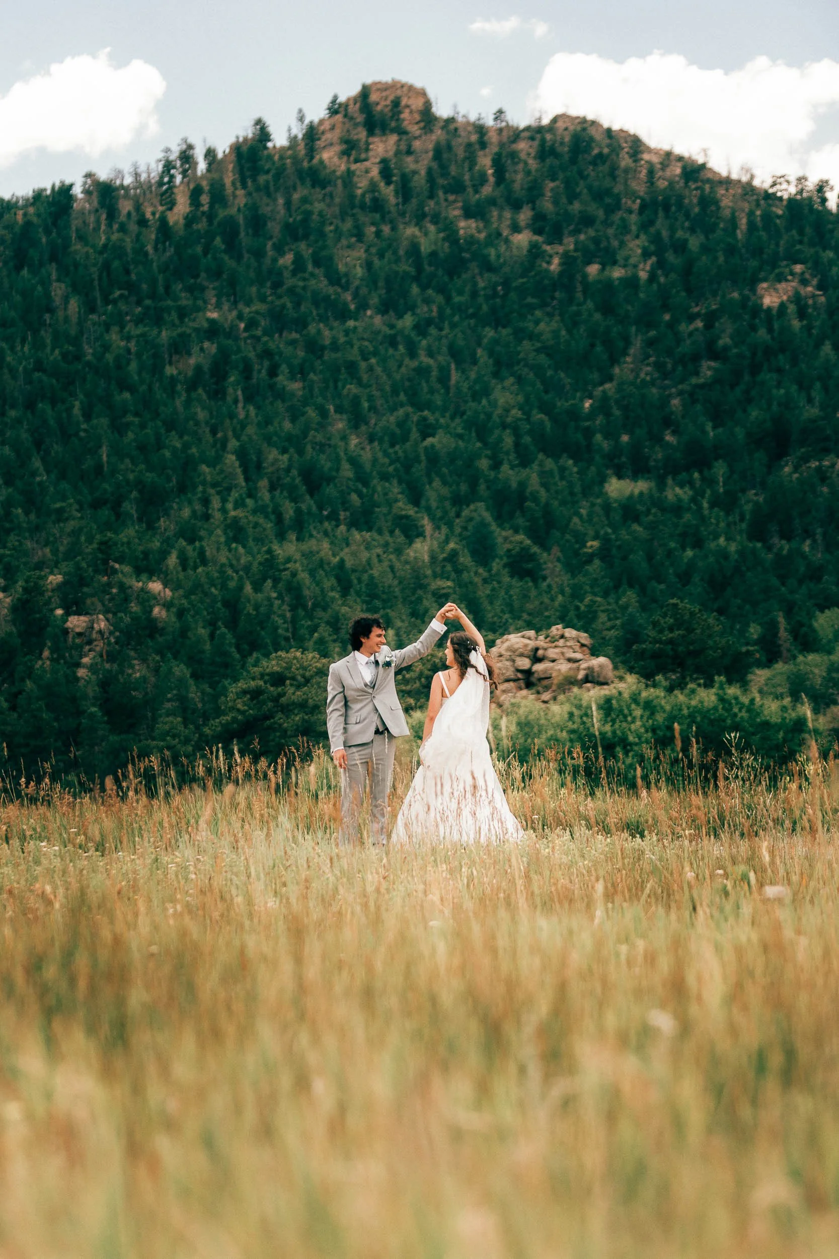 A happy couple holding hands in a field of tall grass against a backdrop of mountains and a cloudy sky posing for a wedding portrait.
