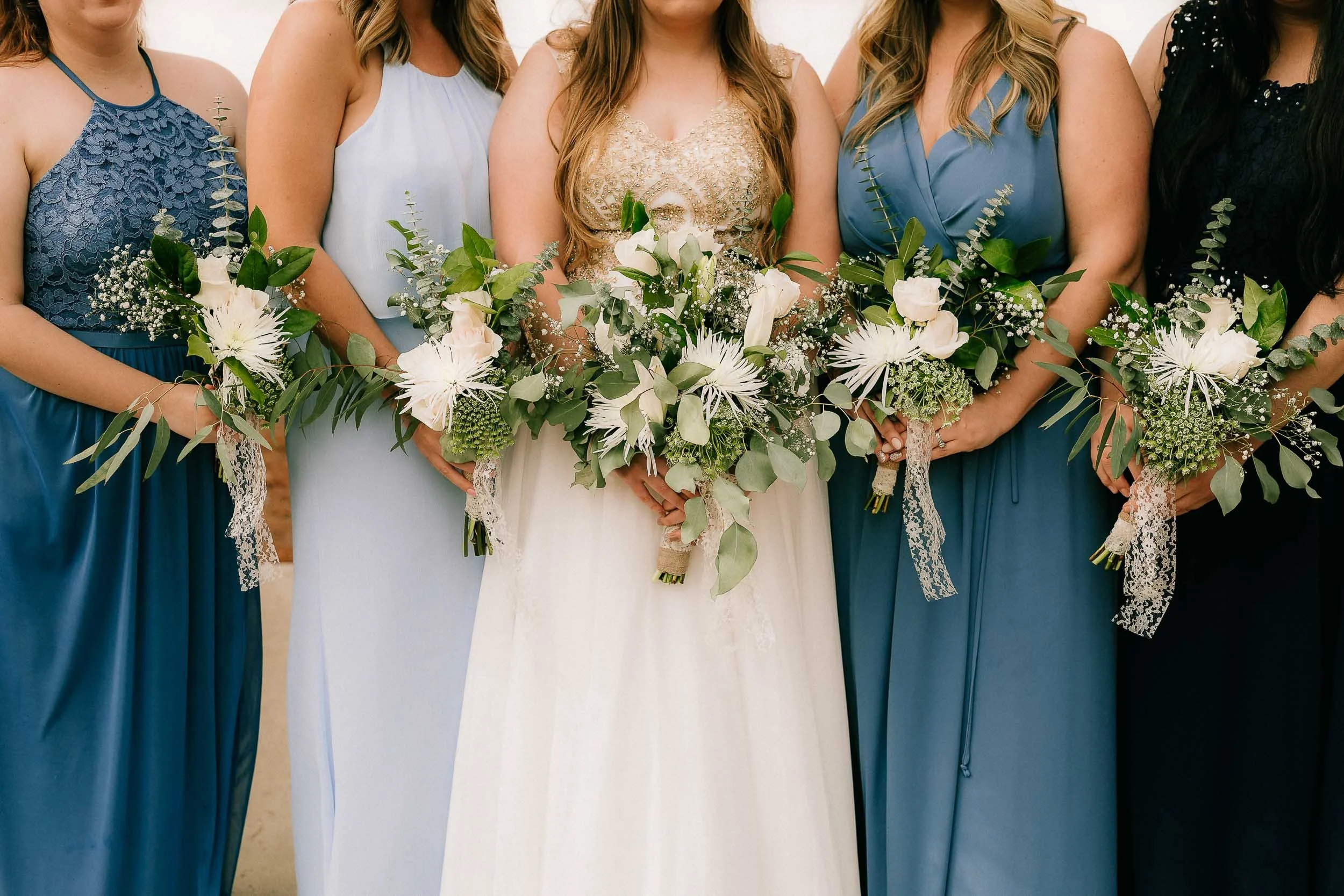 Close-up of bridesmaids holding bouquets at a wedding, with the bride in the center. The bridesmaids are wearing blue dresses, and the bride is wearing a cream-colored dress with intricate gold embroidery.