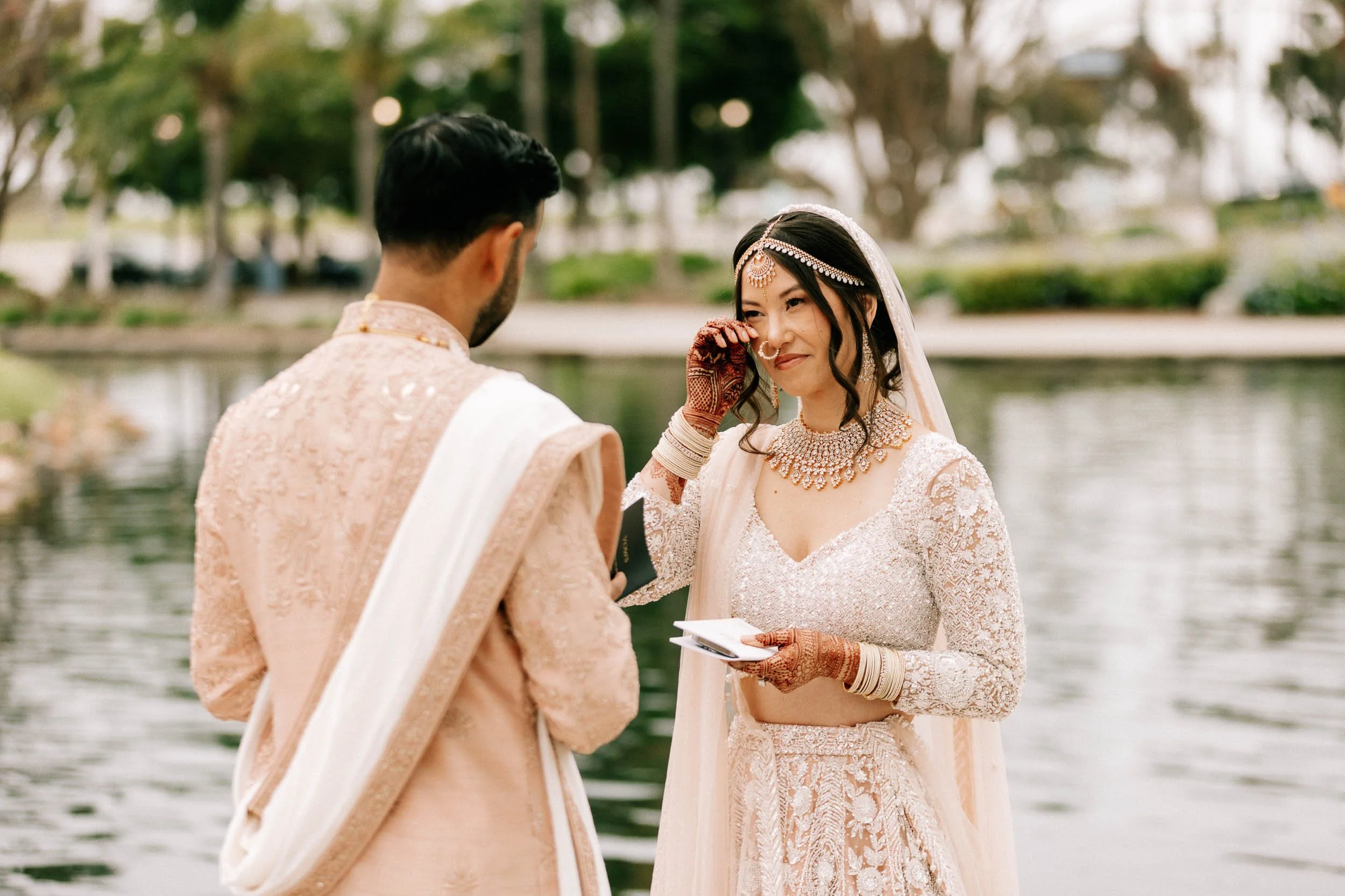 A bride and groom stand by a lake during their wedding ceremony, exchanging vows. The bride is dressed in an elegant lace wedding gown with traditional jewelry, and the groom is in a matching pastel-colored outfit. The bride is wiping away a tear of 