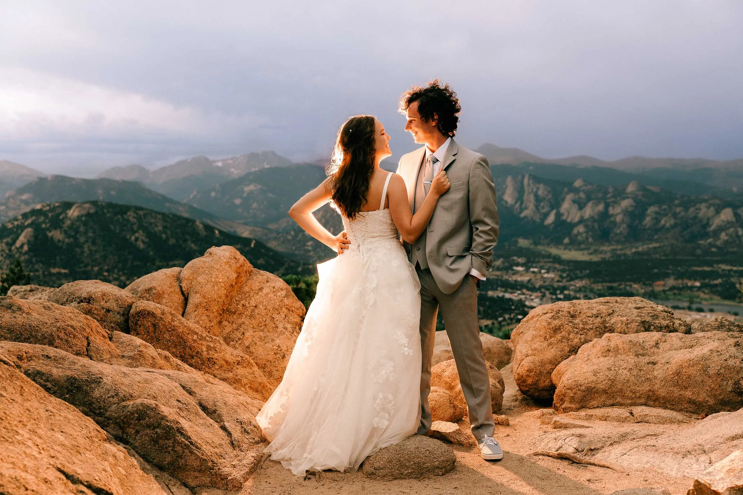 A bride and groom standing on rocks in a mountainous landscape during sunset, looking at each other and smiling.