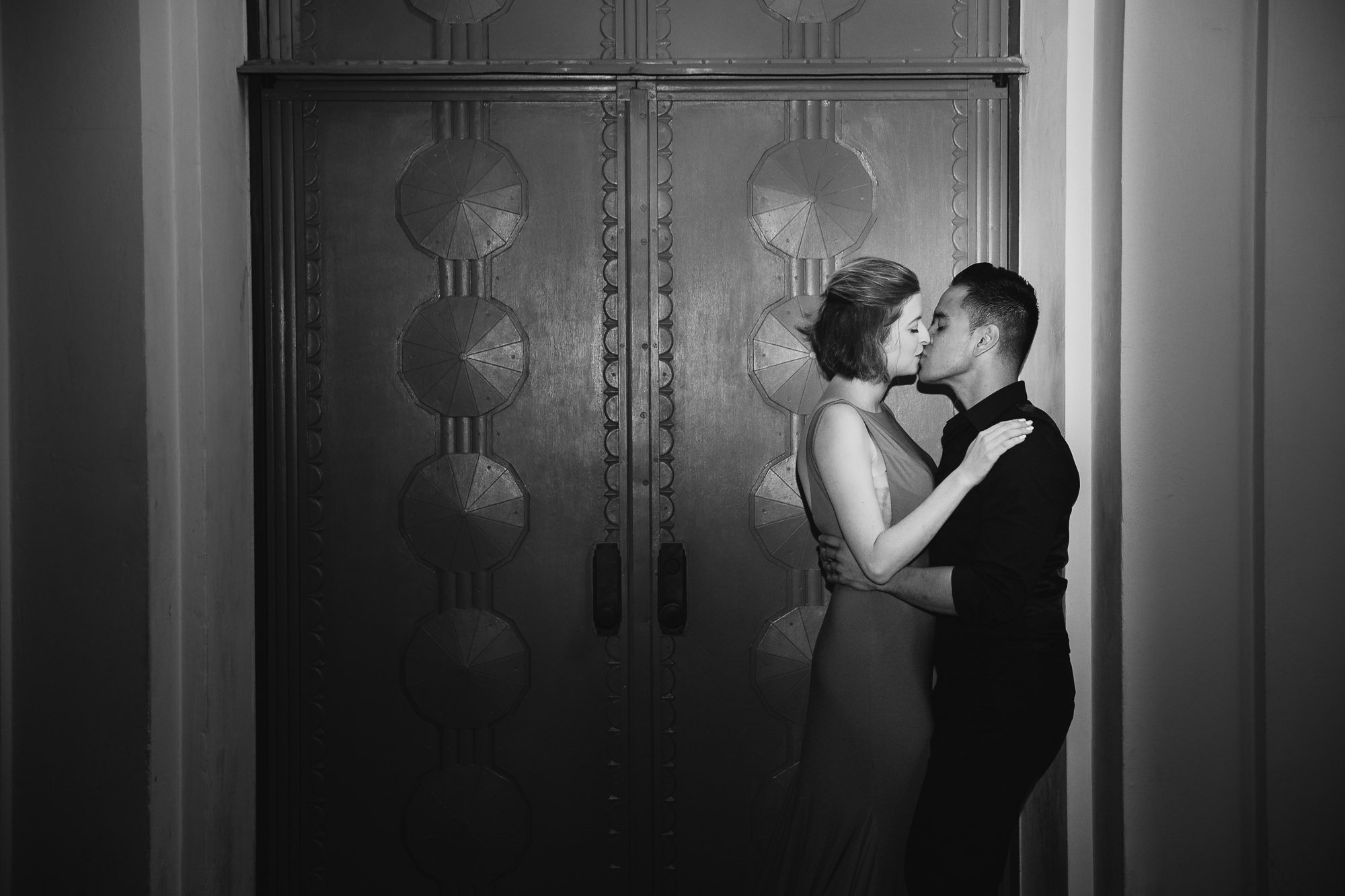 A black-and-white photo of a couple kissing in front of large double doors with ornate designs. The woman has short hair and is wearing a sleeveless dress, while the man is wearing a dark shirt.