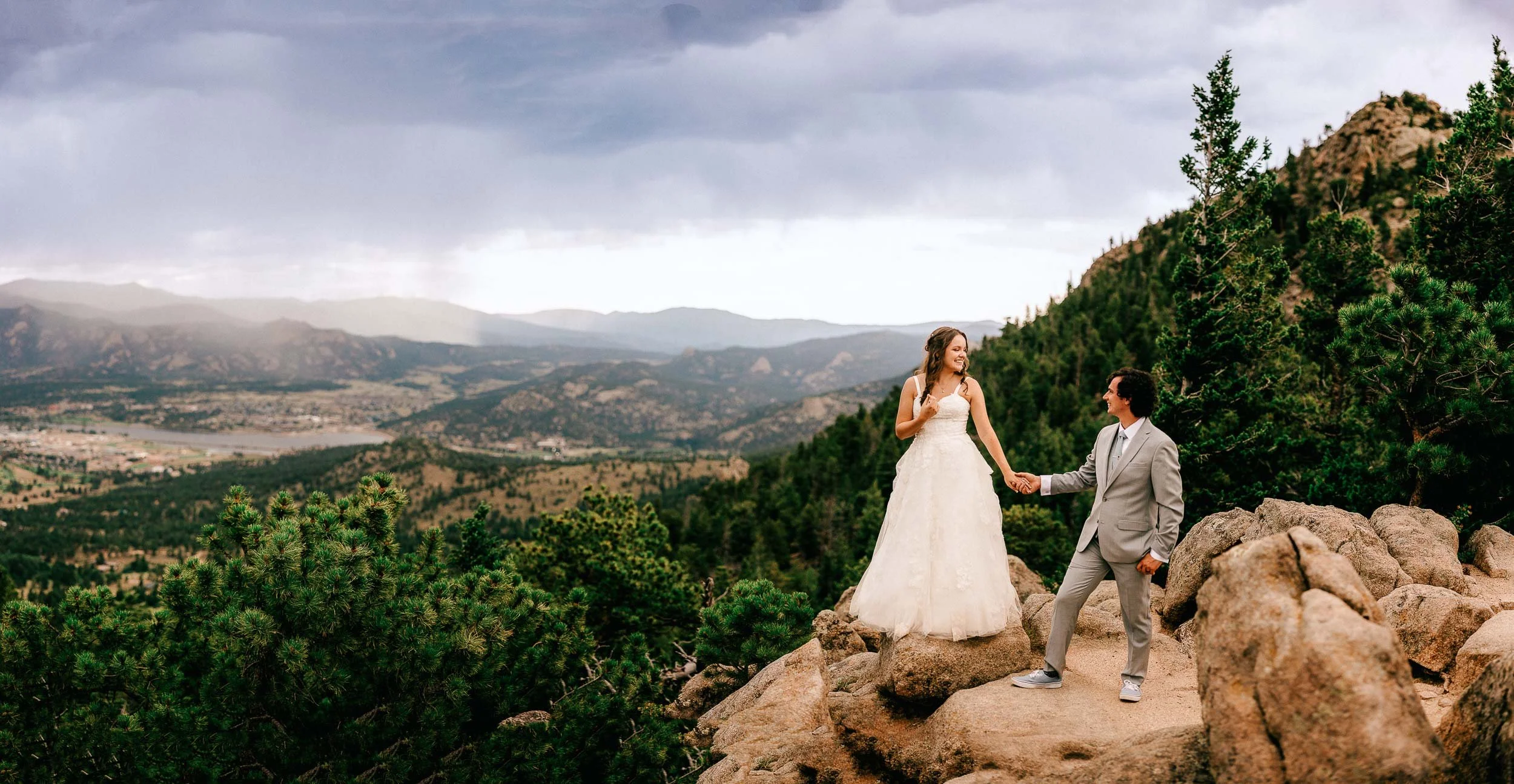 A bride and groom standing on rocks outdoors with a mountain and cloudy sky background, holding hands and smiling at each other.