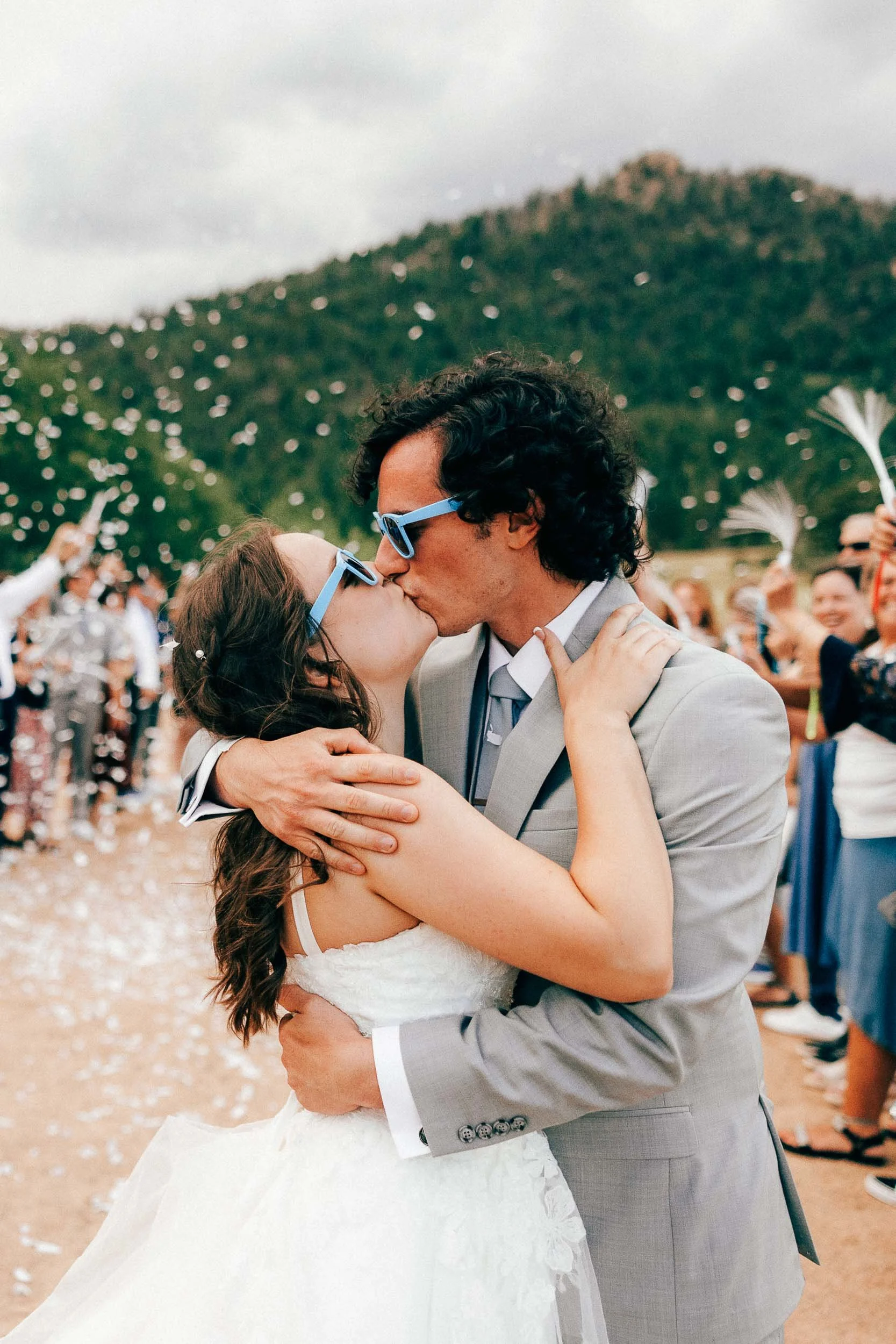 A bride and groom kiss during their wedding ceremony outdoors, with guests watching and throwing flower petals in the background.