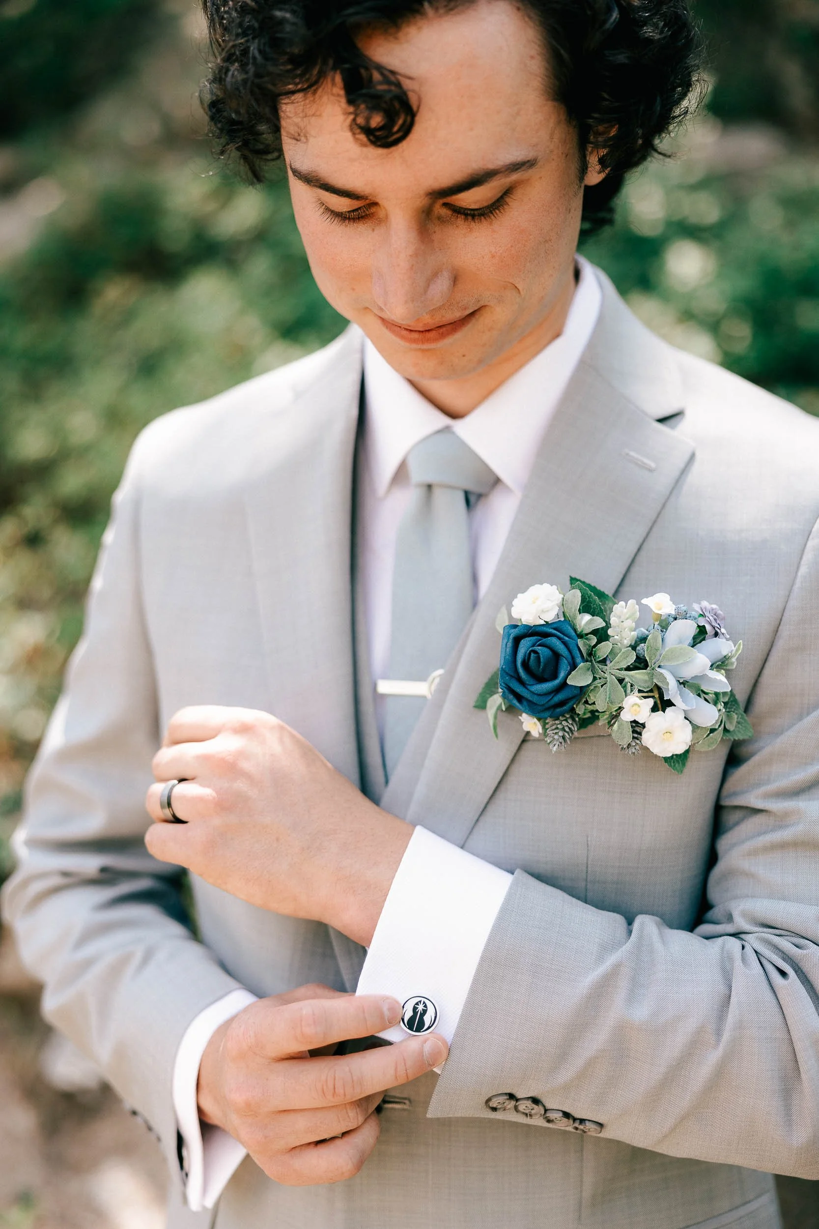 Close-up of a man in a light gray suit adjusting his cufflink, wearing a boutonnière with blue and white flowers, with a blurred outdoor background on his wedding day.