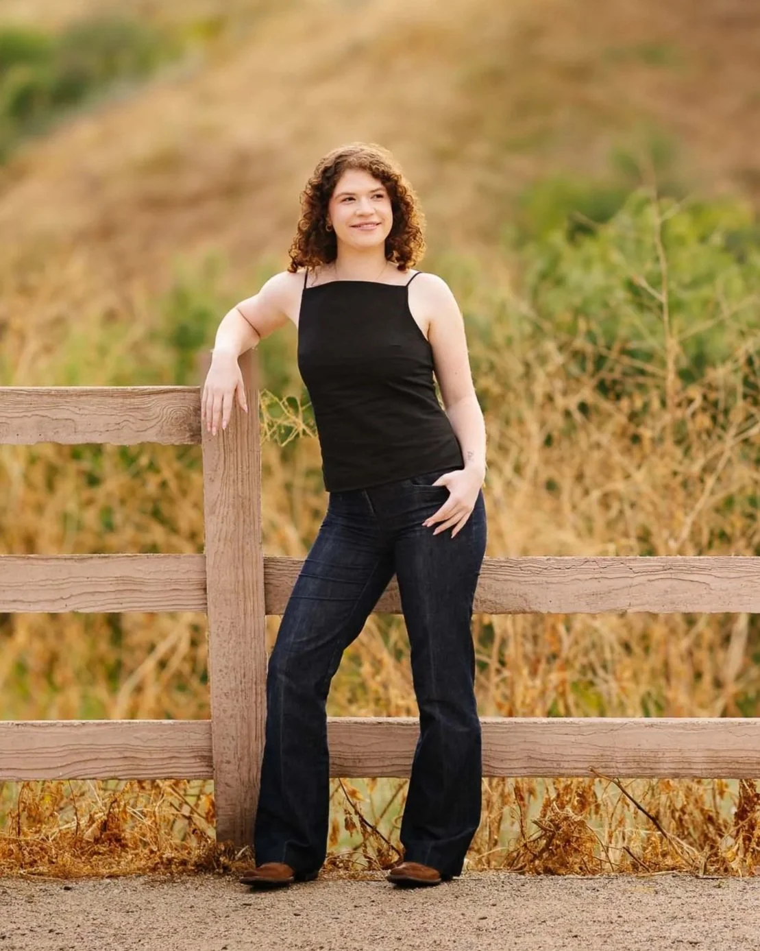 a caucasin curly haired girl posing for senior portraits in a field with a fence and black shirt and blue jeans