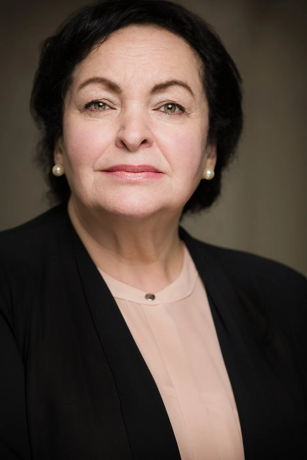 Portrait of a middle-aged woman with short dark hair, wearing pearl earrings, a light pink blouse, and a black blazer against a neutral background.