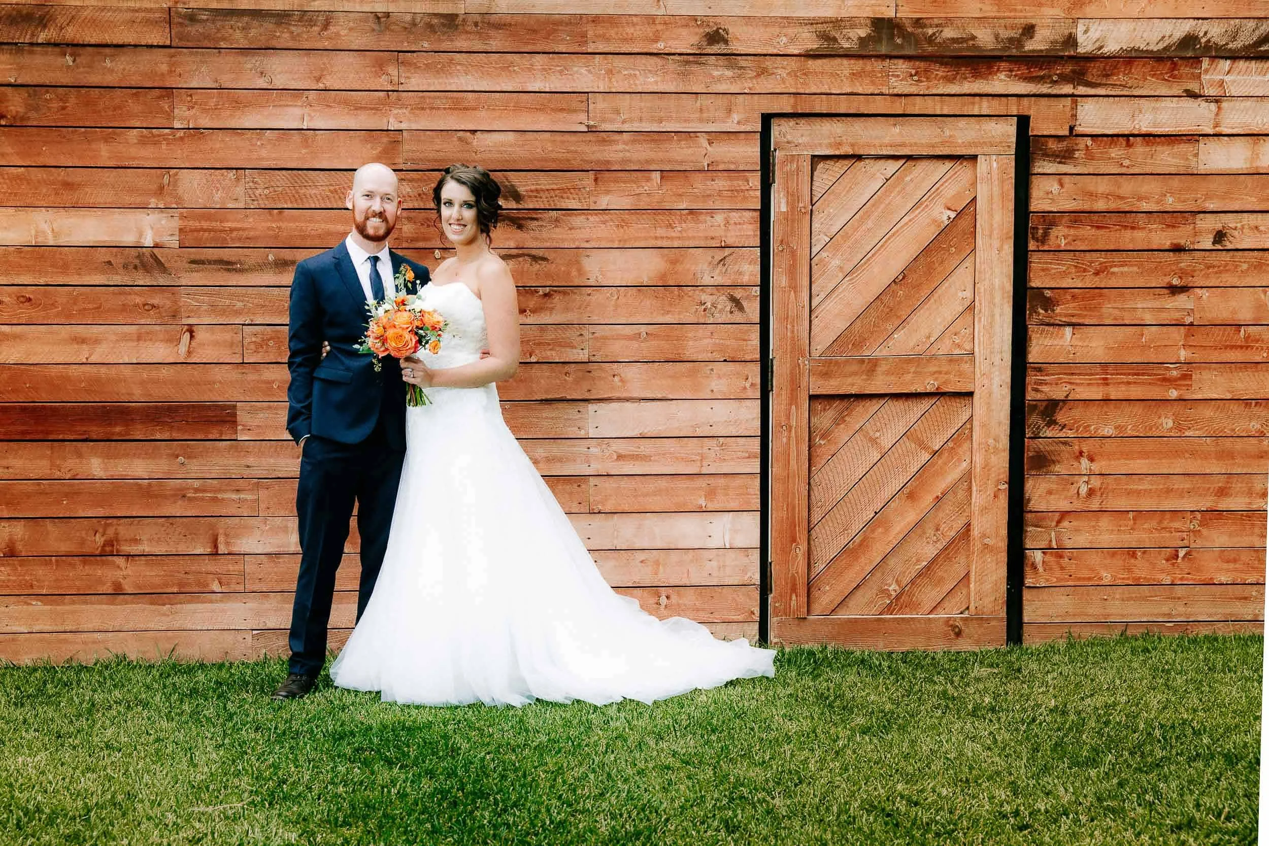 A newlywed couple standing on grass in front of a large wooden barn wall, smiling. The groom is in a navy suit holding a bouquet, and the bride is in a white wedding dress.