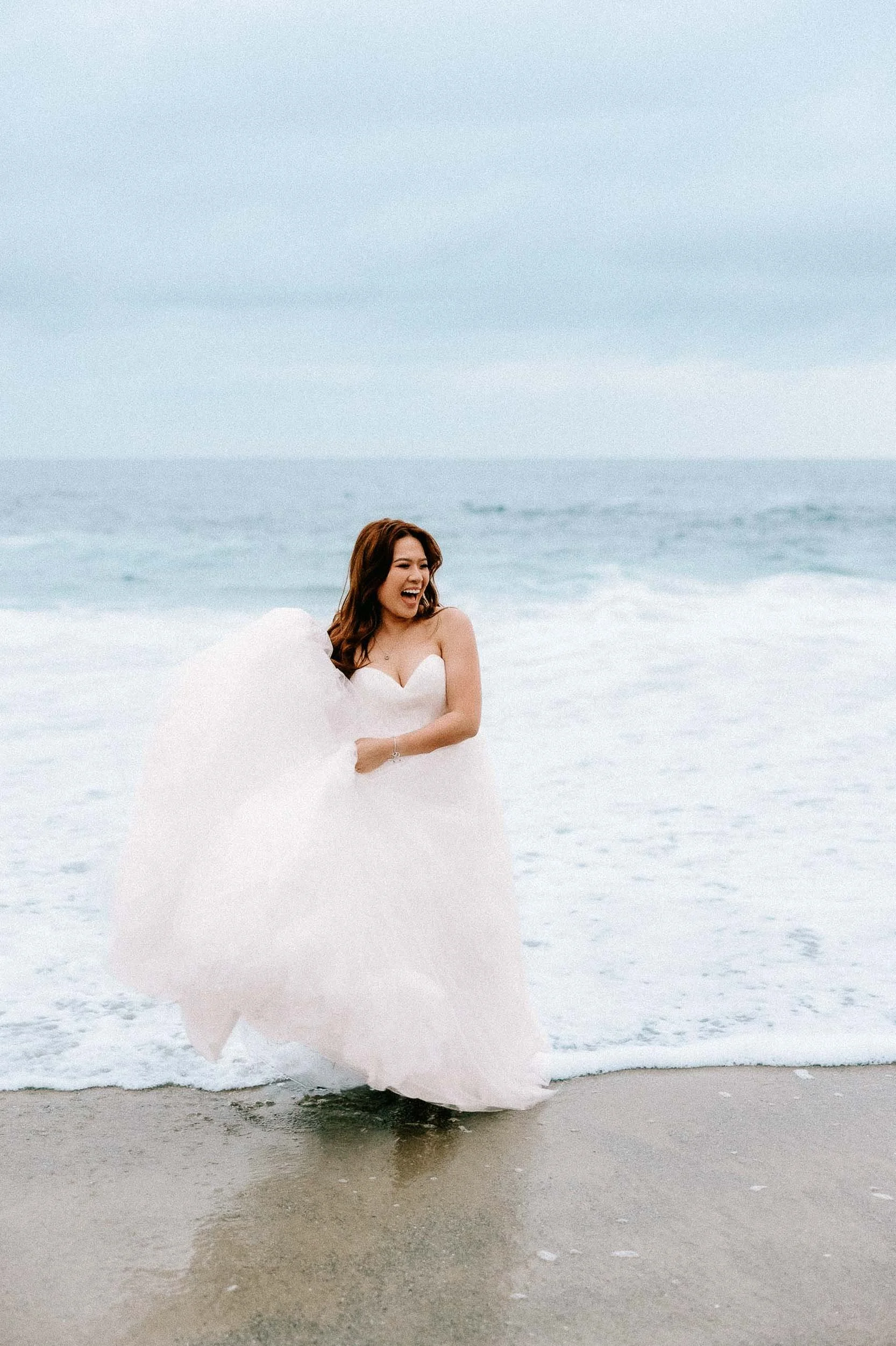 A woman in a white wedding dress standing on the beach with ocean waves behind her, laughing and holding the dress with her hands.