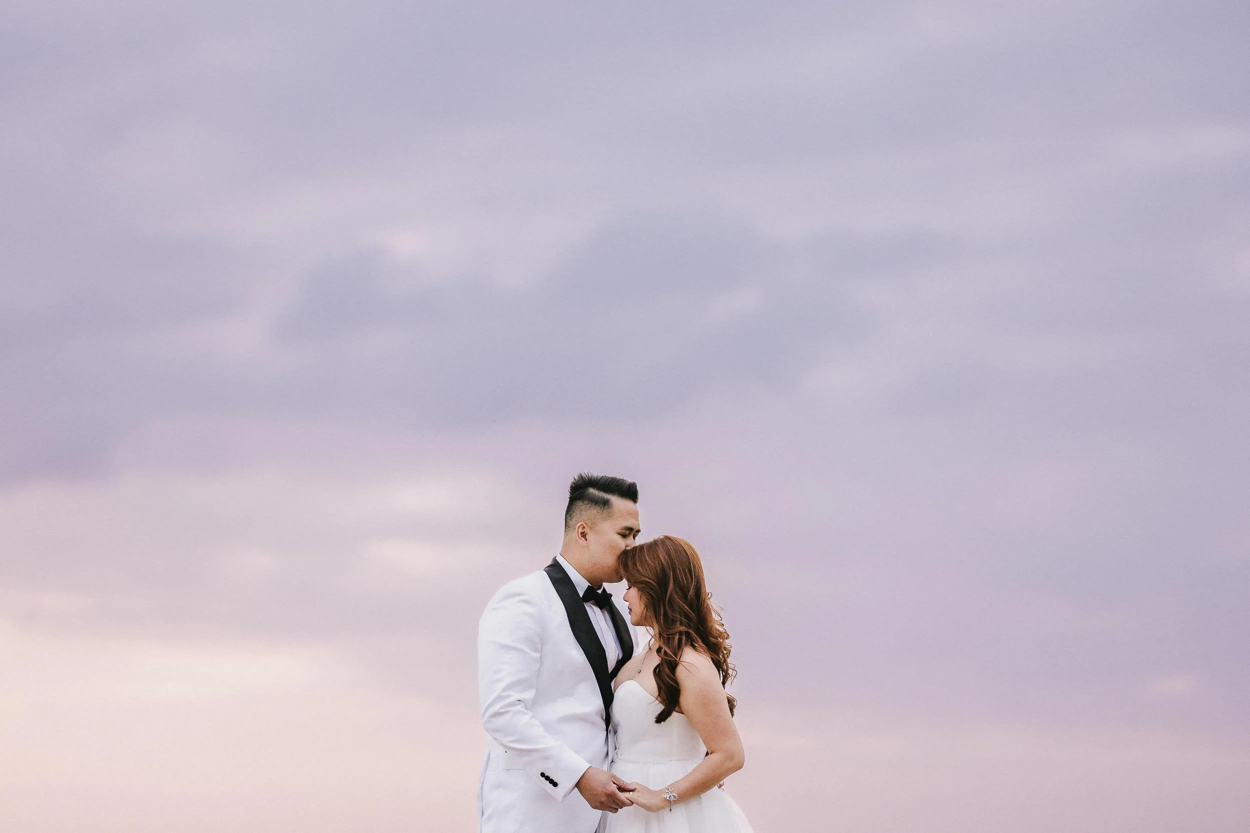 A bride and groom in wedding attire share a kiss under a cloudy pink sky, holding hands.