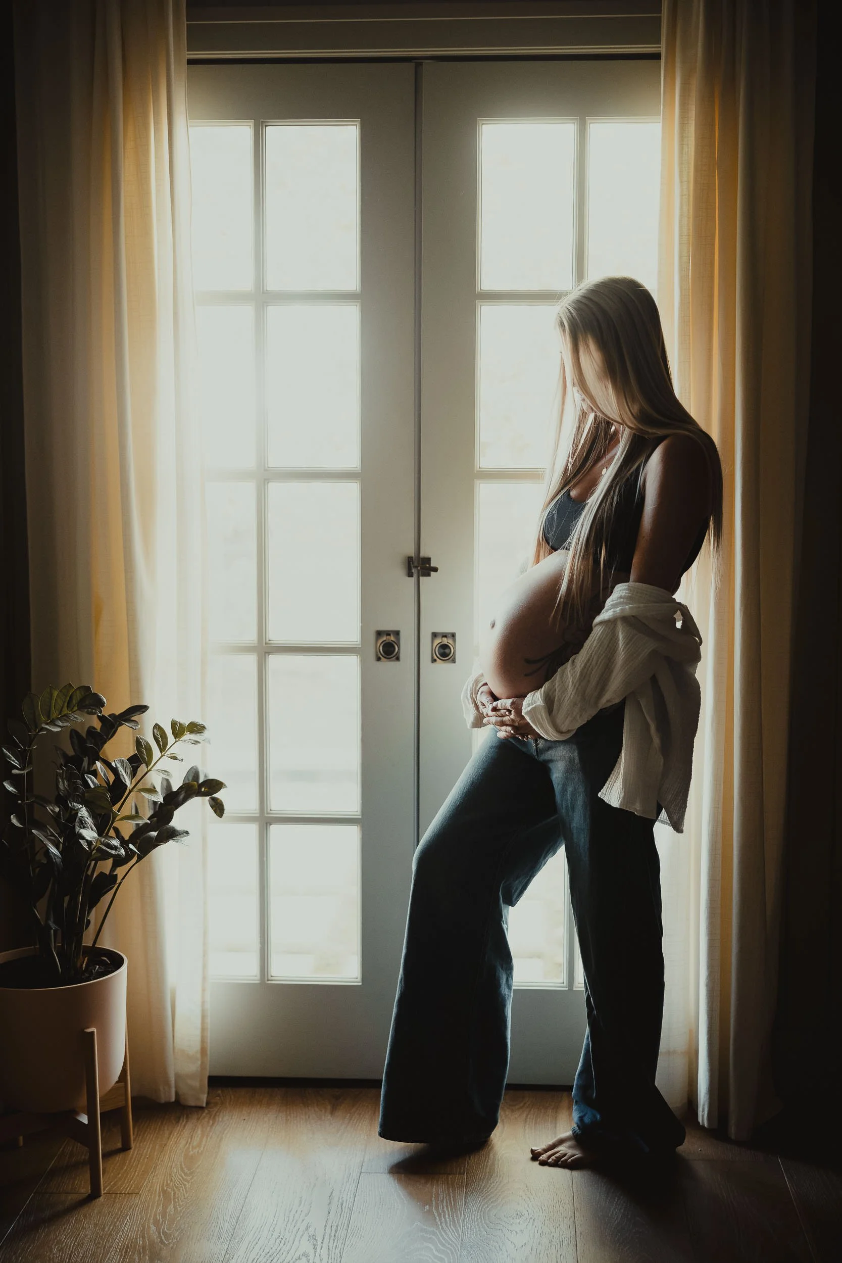 A woman with long blonde hair stands barefoot in front of a glass-paneled door, backlit by natural light. She is wearing a black top and jeans, and a light-colored shirt is draped over her shoulders. To the left, there is a potted plant with lush green leaves, and sheer curtains frame the door.