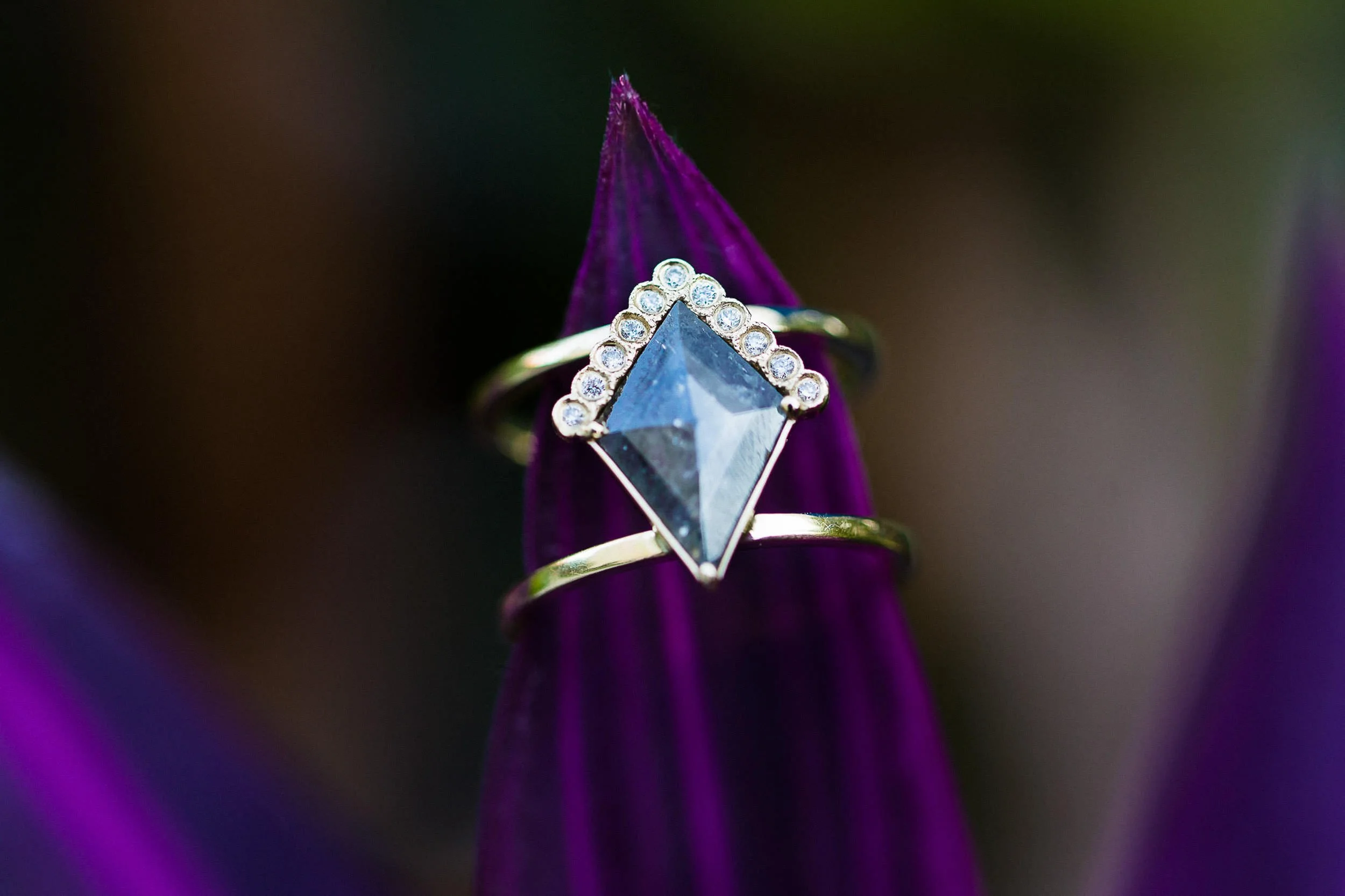 Close-up of an engagement ring with a large geometric blue gemstone surrounded by small diamonds, worn on a finger with a purple flower petal in the background.