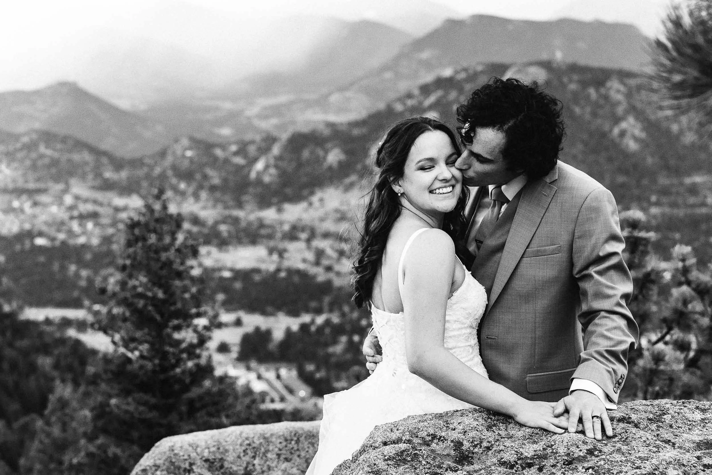 Black and white photo of a couple in wedding attire, standing close together outdoors, with mountains and a landscape in the background. The man is kissing the woman's forehead, and they are holding hands.