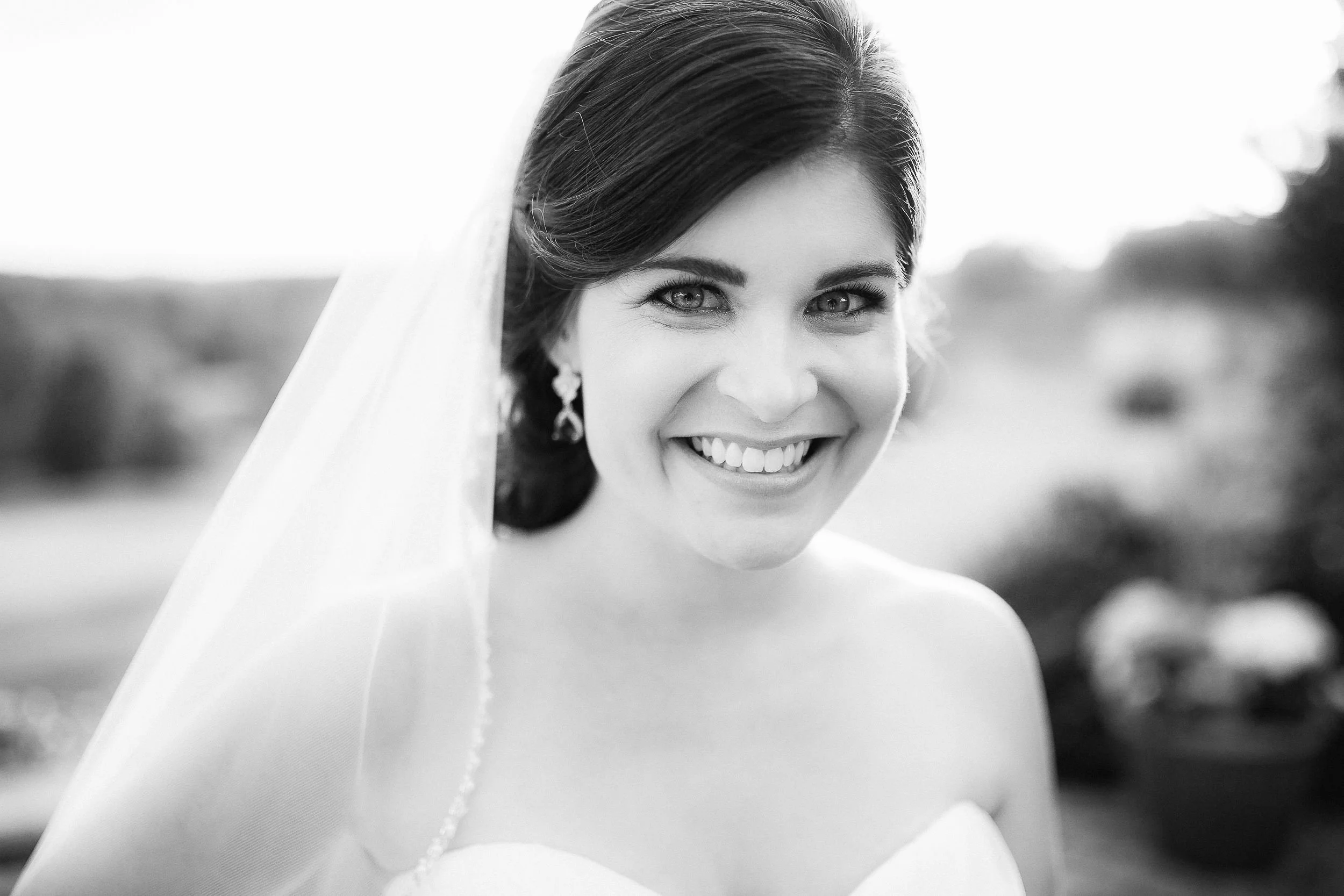 Black and white portrait of a smiling woman with dark hair, earrings, and a radiant expression outdoors.