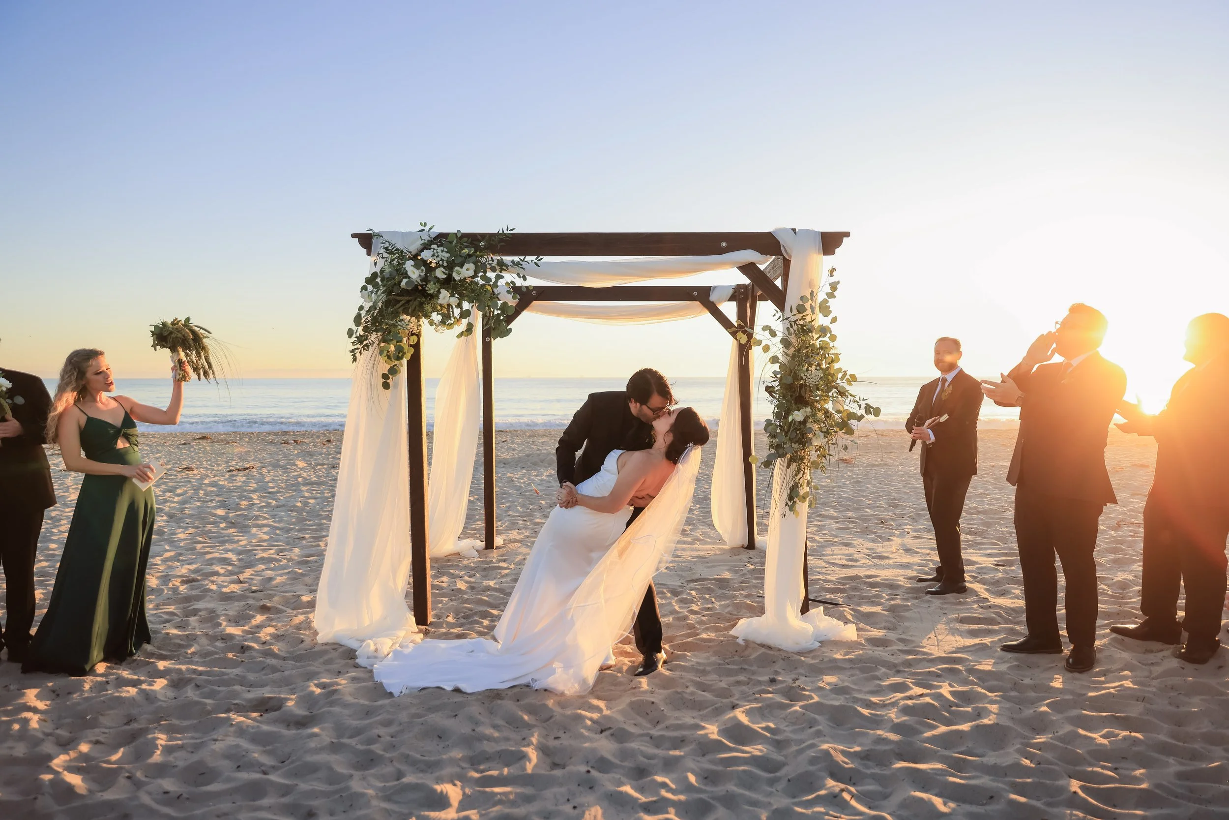 A wedding ceremony taking place on a beach at sunset, featuring a couple kissing under a decorated wooden arch, surrounded by friends and family.