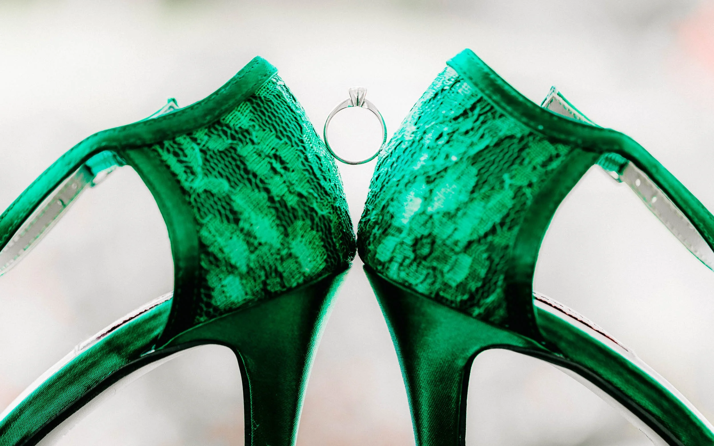 Green high-heeled shoes with a wedding ring placed between them, photographed from a low angle against a white background.
