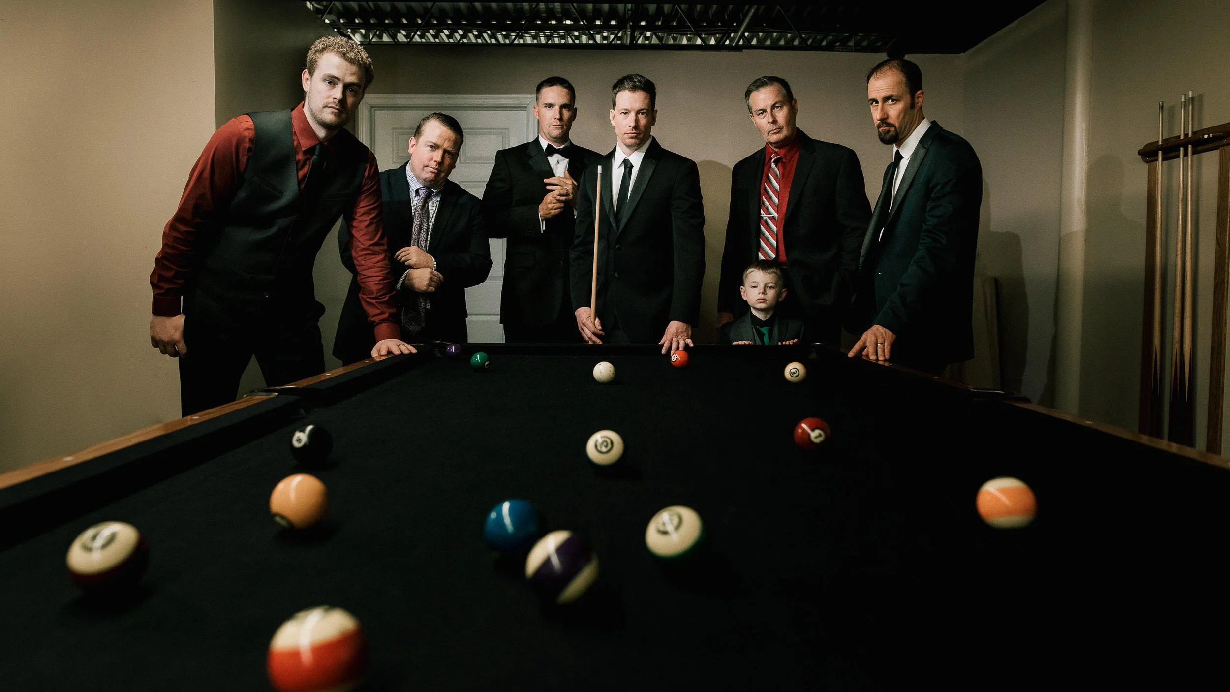 Seven men in suits and a boy in a tuxedo standing around a pool table with billiard balls, in an indoor game room.