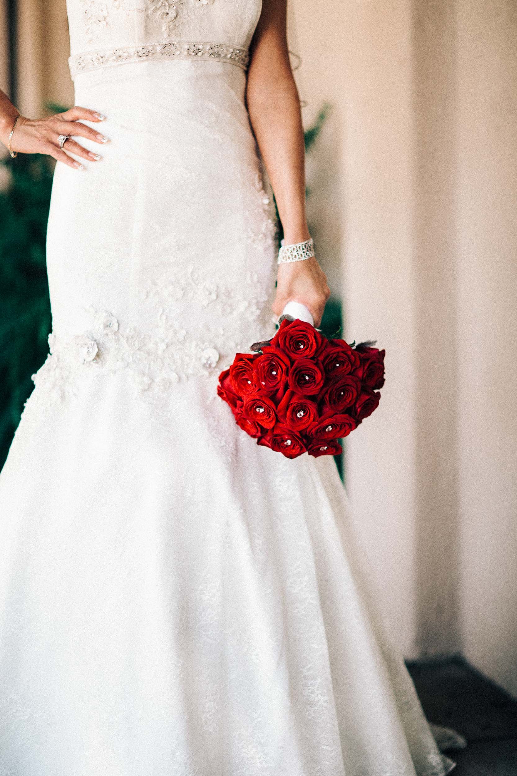 A bride in a white wedding dress holding a bouquet of red roses. The dress has lace details and she is wearing jewelry, including a bracelet and rings.