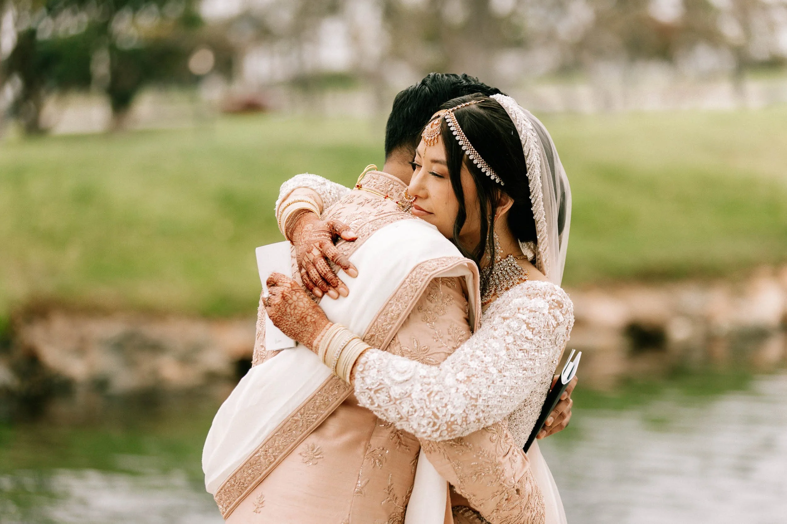 A bride and groom embrace outdoors on their wedding day, with the bride wearing traditional jewelry and a lace dress, and the groom in a cream-colored traditional outfit, blurred greenery background.
