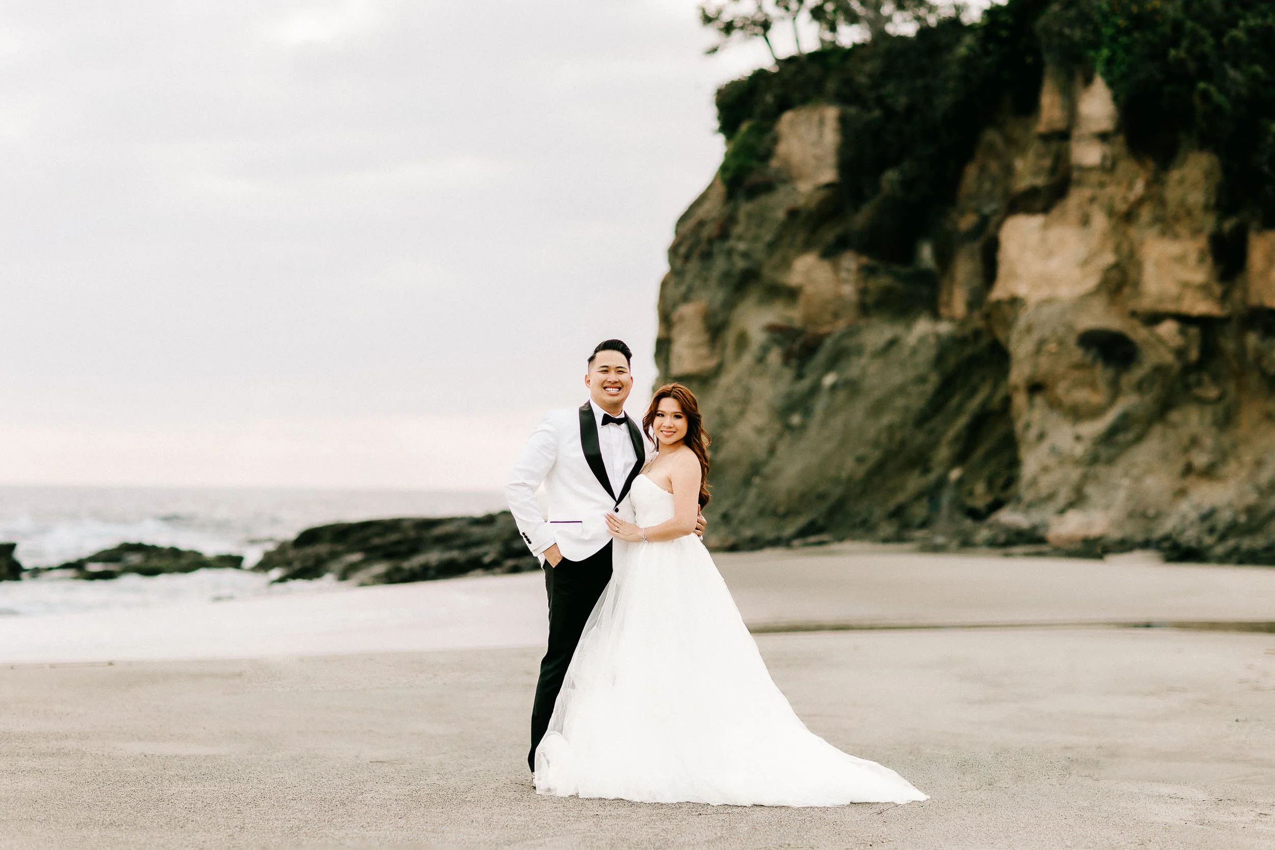 A happy couple in wedding attire standing on a sandy beach with cliffs in the background, during overcast weather.