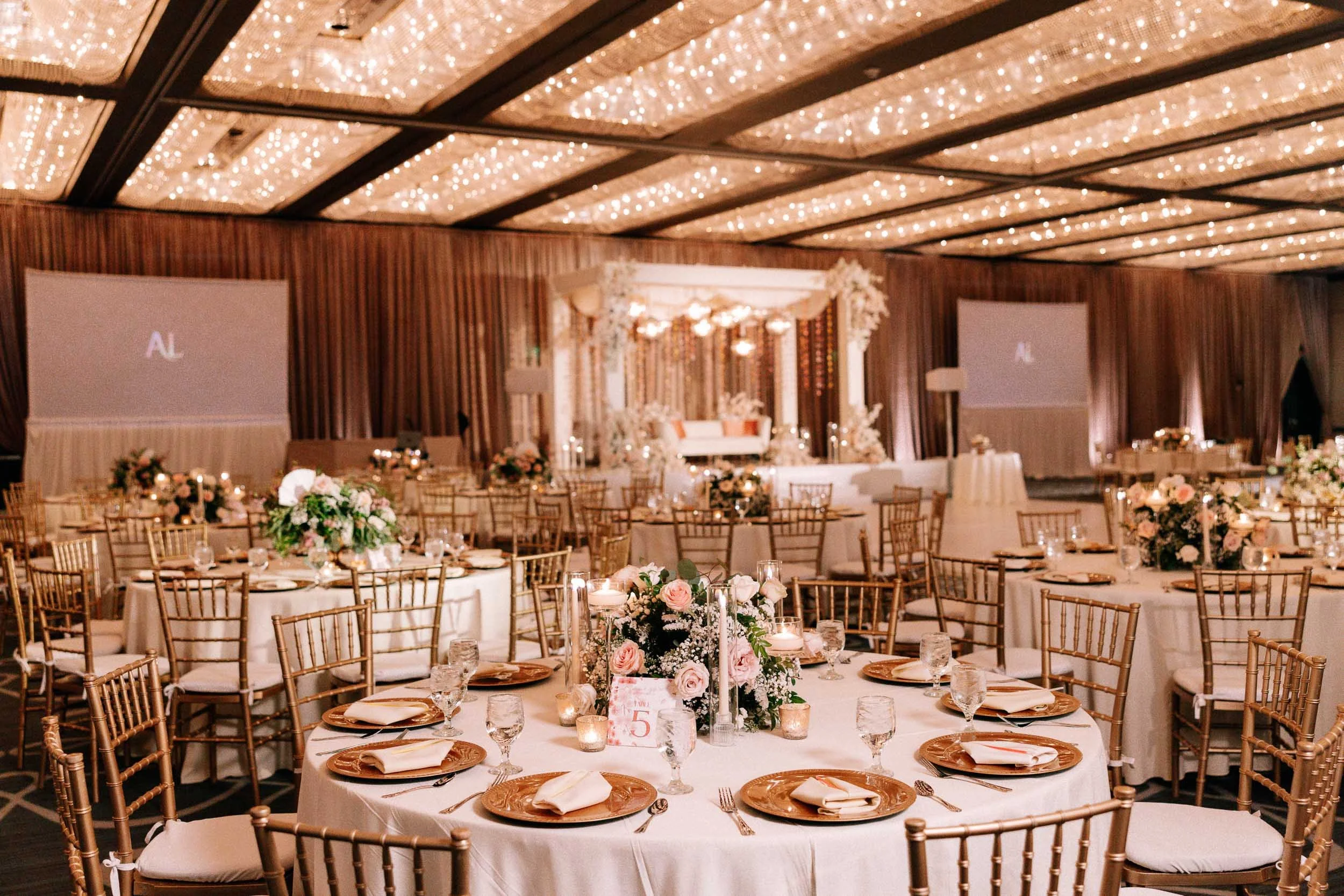 Elegant wedding reception setup with round tables, white tablecloths, floral centerpieces, gold chairs, and a decorated stage with fairy lights on the ceiling.
