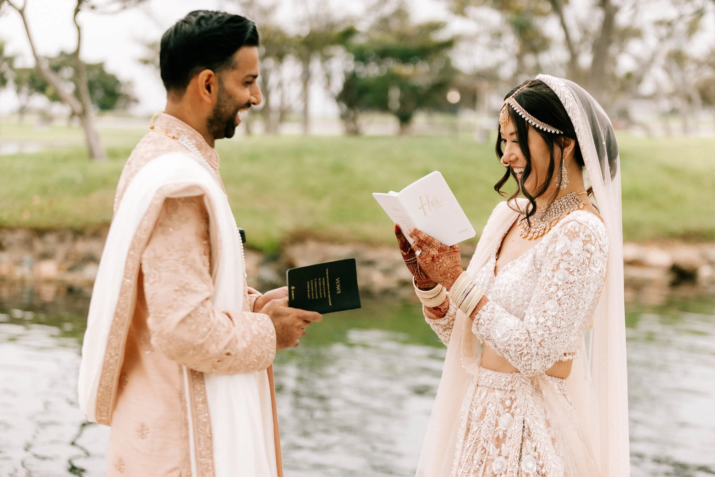 A bride and groom are exchanging vows outdoors near a water body, with trees in the background. They are dressed in traditional wedding attire; the bride is wearing a white lace dress with jewelry, and the groom is in a light-colored outfit. They are
