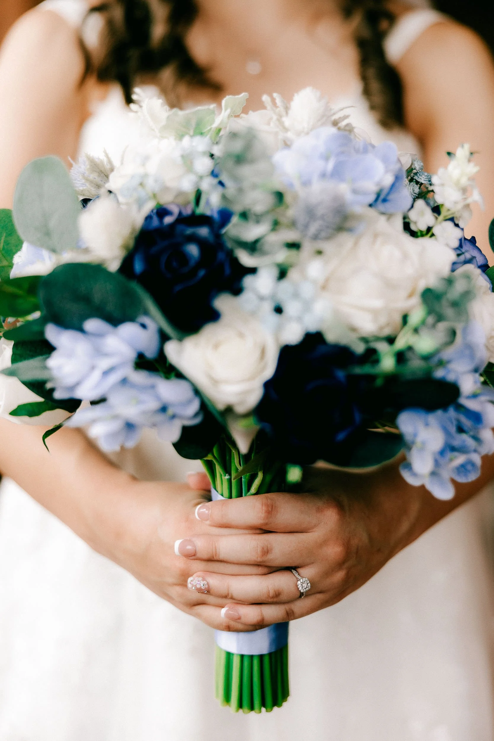 Close-up of a bride holding a bouquet of white, blue, and purple flowers with greenery, showing her hands with wedding rings.