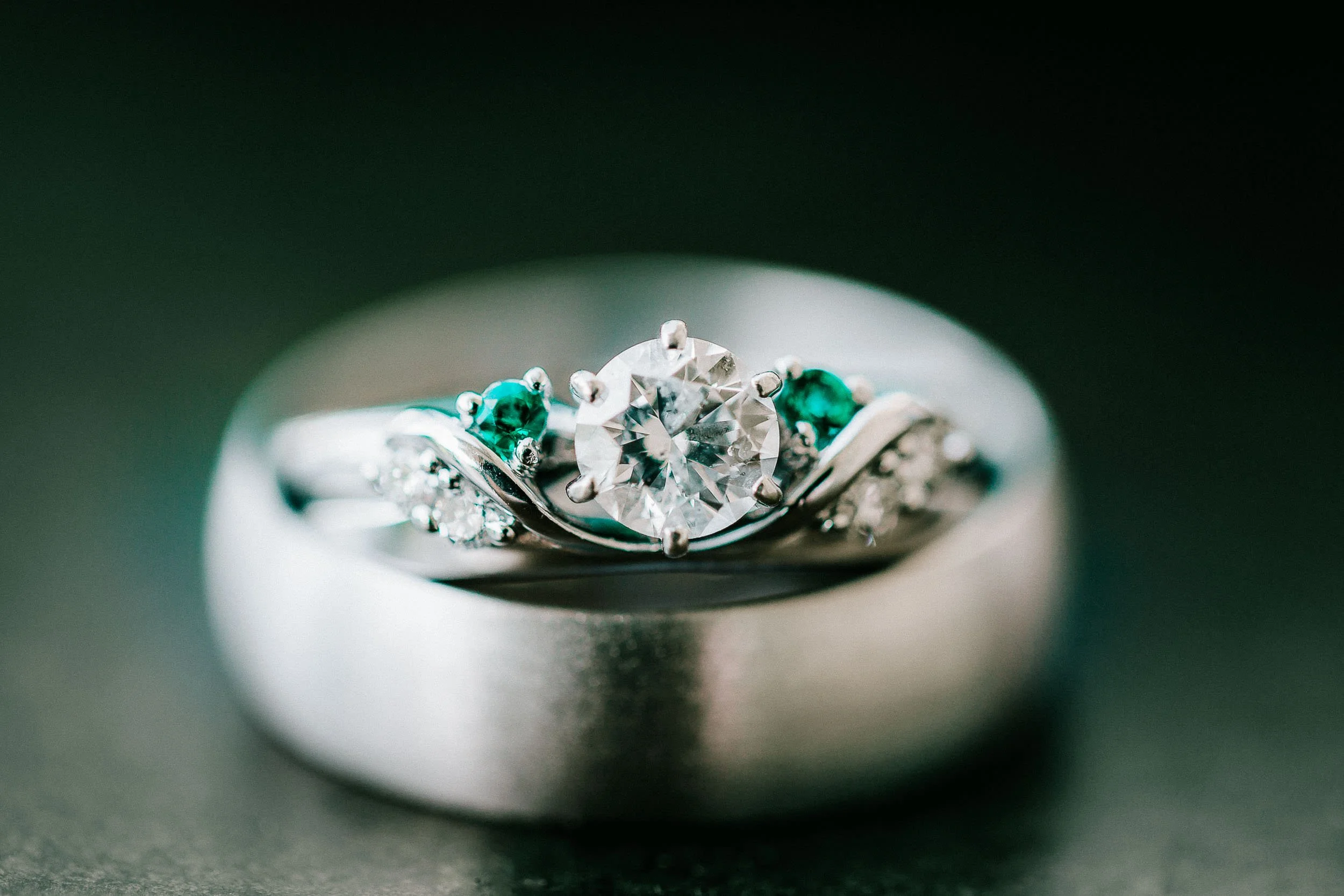 Close-up wedding photo of a silver ring with a large central clear diamond, flanked by smaller emerald and diamond stones.