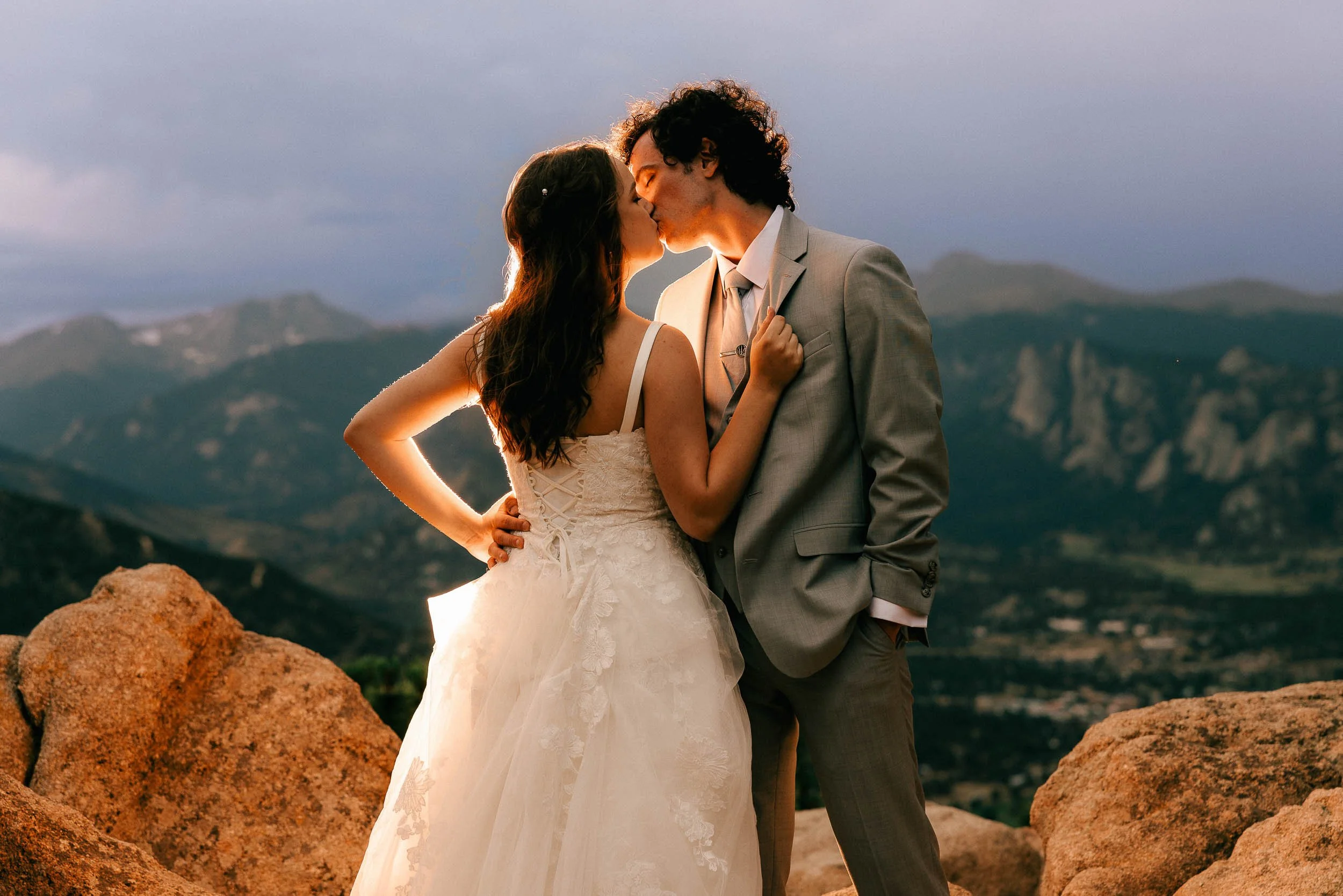 A bride and groom sharing a kiss outdoors during sunset, with mountains in the background.