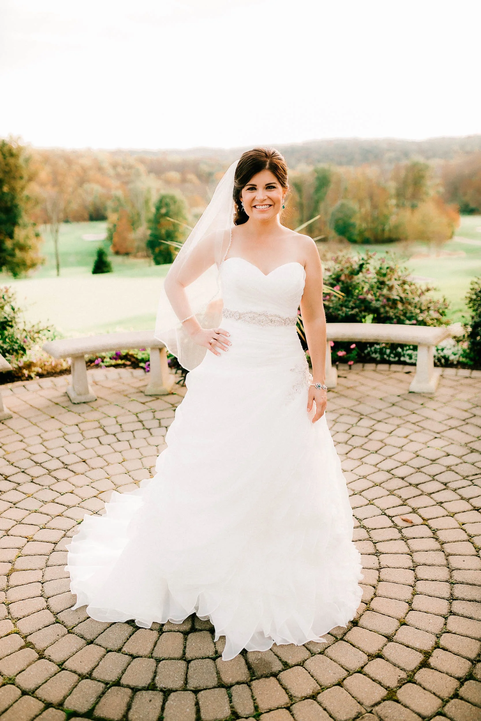 A bride with dark hair in an updo, wearing a strapless white wedding gown with a beaded waistband, standing outdoors on a cobblestone path with a scenic background of green fields, trees, and a distant hill. She is smiling, with a veil hanging from h