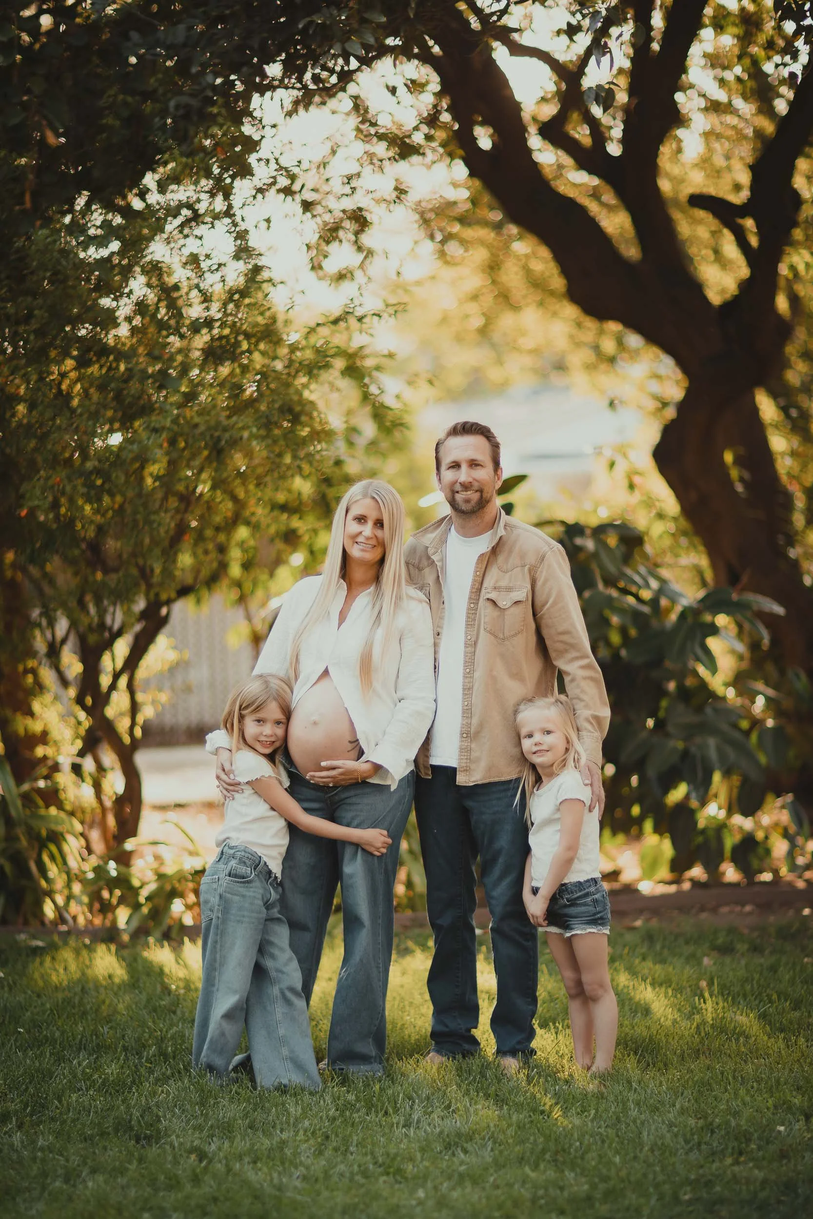 A family photo taken outdoors during sunset, featuring a pregnant woman, her partner, and their two children standing on a grassy area with trees in the background.