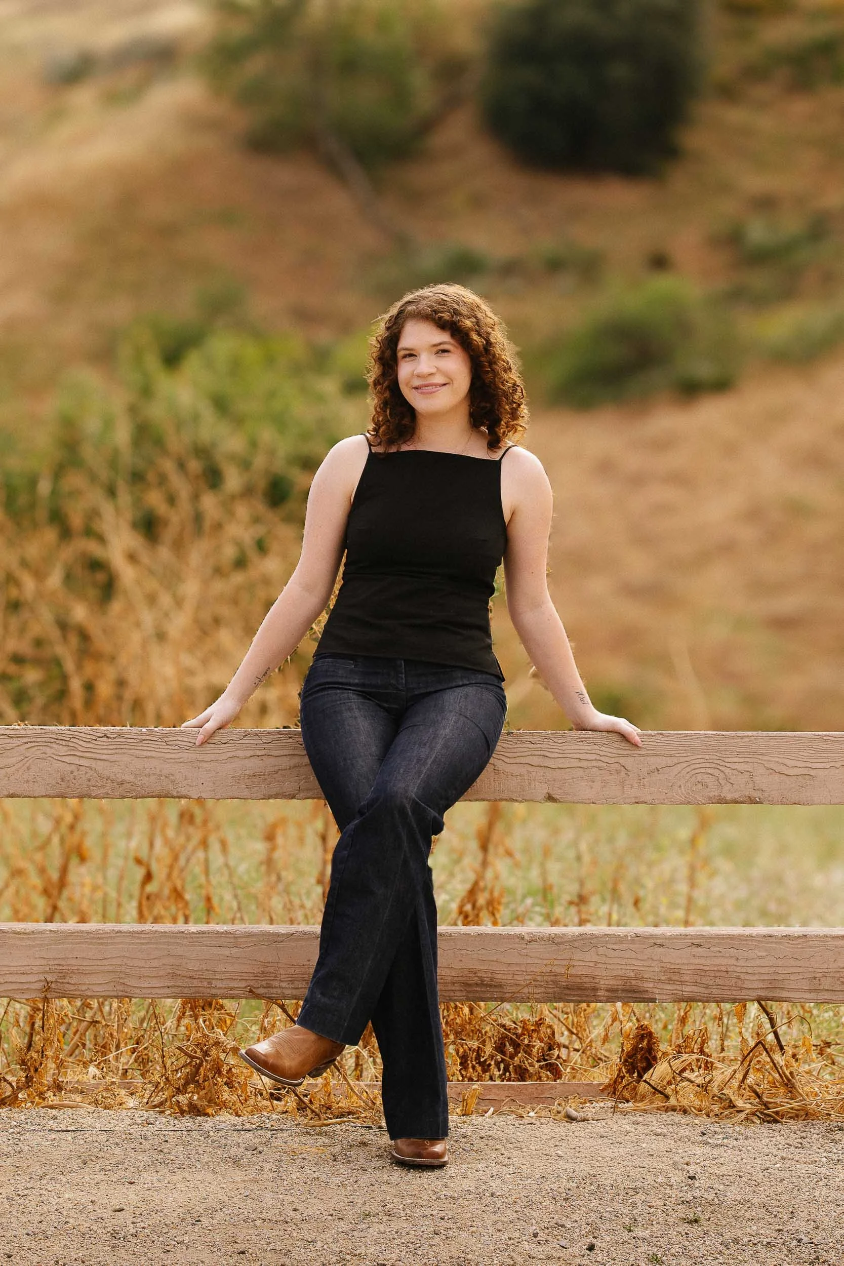 A woman with curly brown hair sitting on a wooden fence in a rural outdoor setting during autumn, wearing a black sleeveless top, dark jeans, and brown shoes.