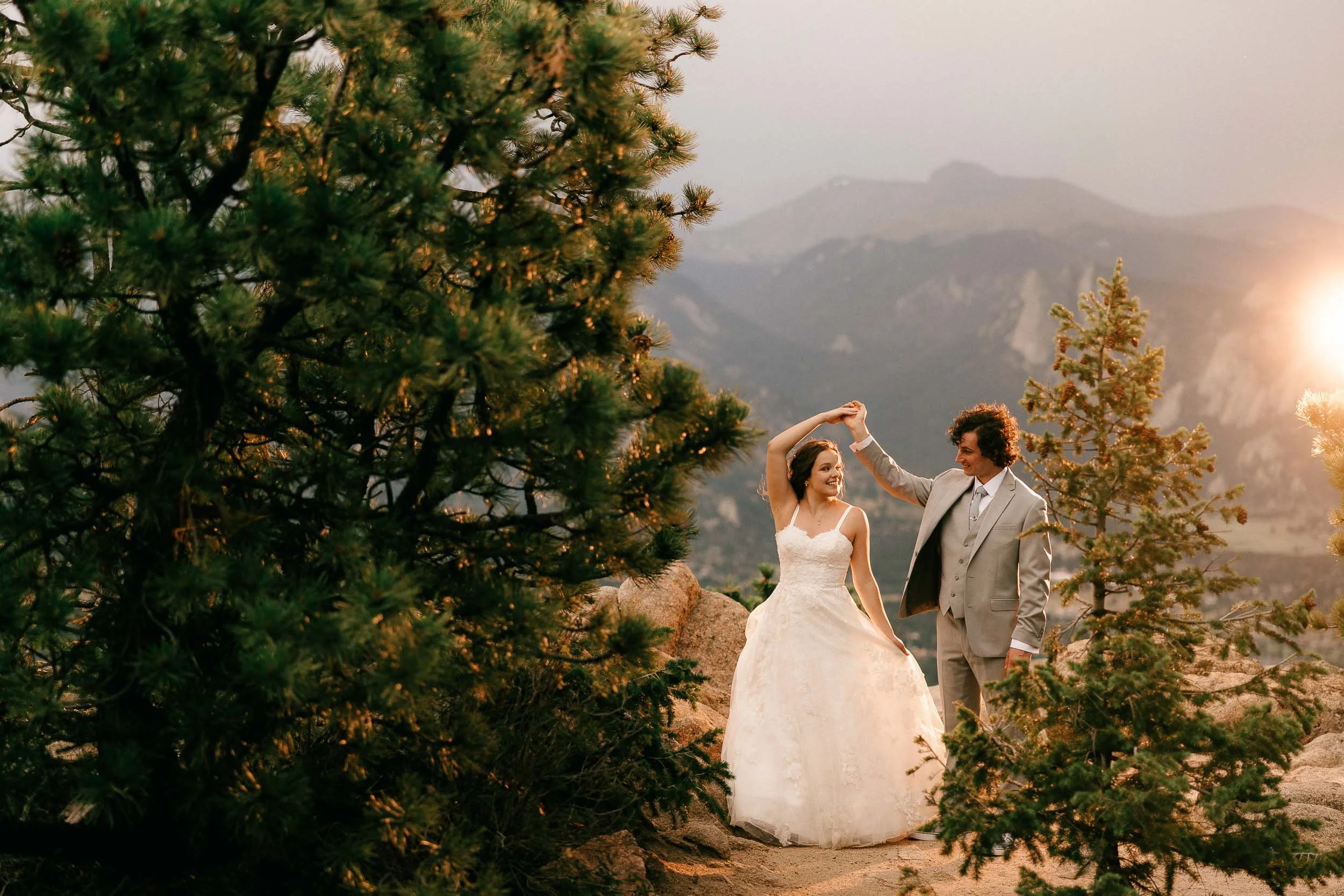A bride and groom dancing outdoors during sunset, surrounded by trees and mountains in the background.