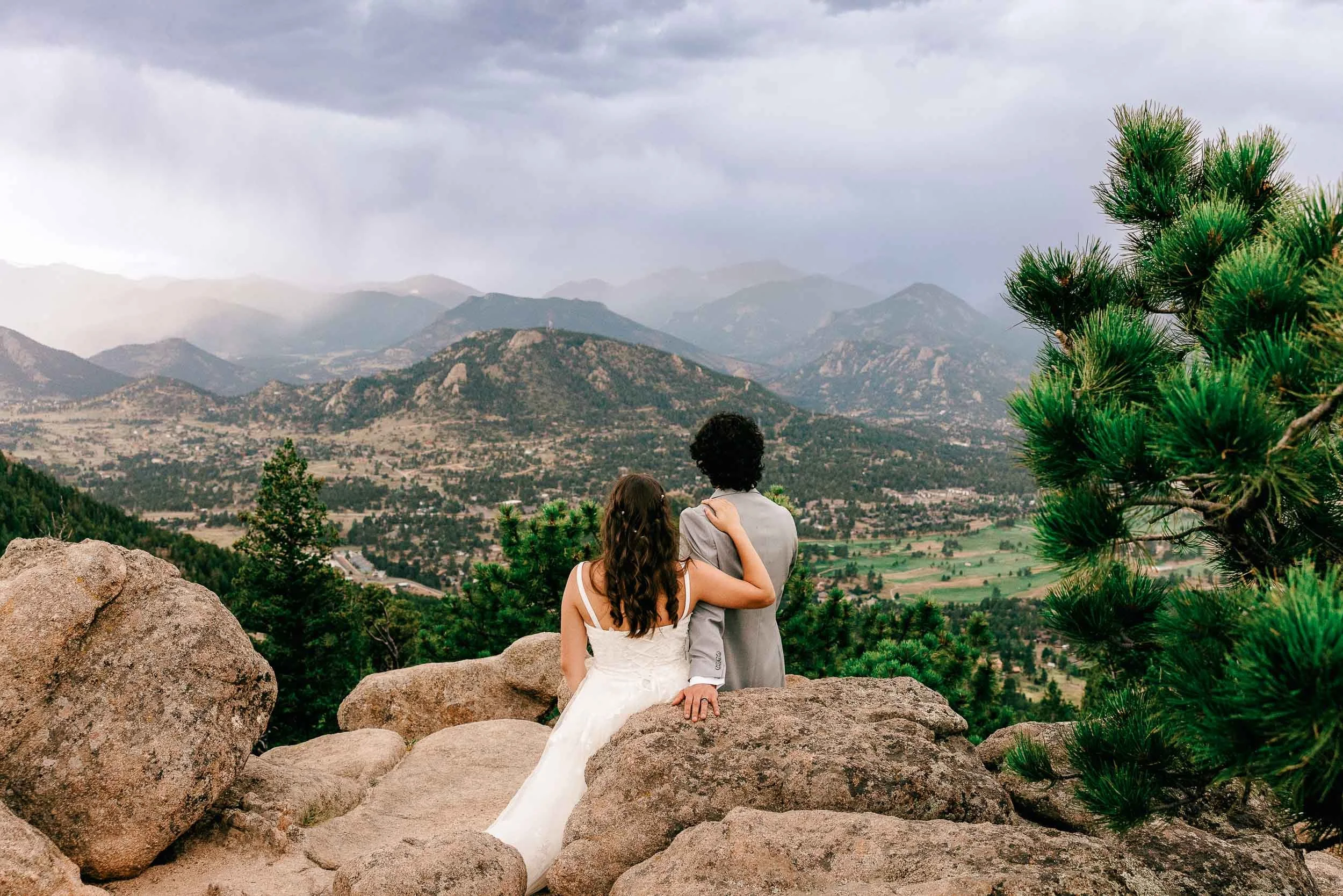 A couple sitting on rocks in front of a mountain landscape, with the woman in a white wedding dress and the man in a gray suit, embracing each other, with lush green trees and cloudy sky on their wedding day.