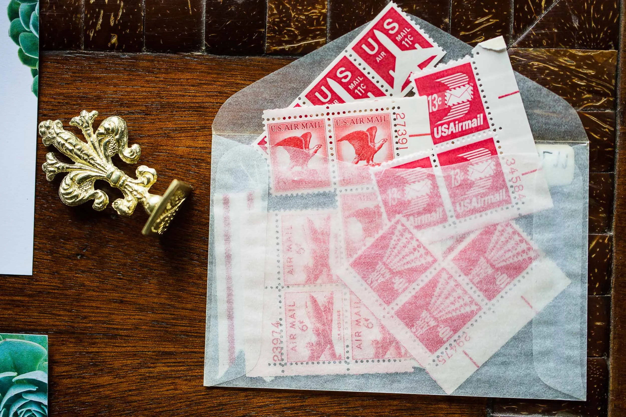 A bunch of red and white U.S. Air Mail postage stamps on a piece of translucent paper, placed on a wooden surface with a decorative golden element.