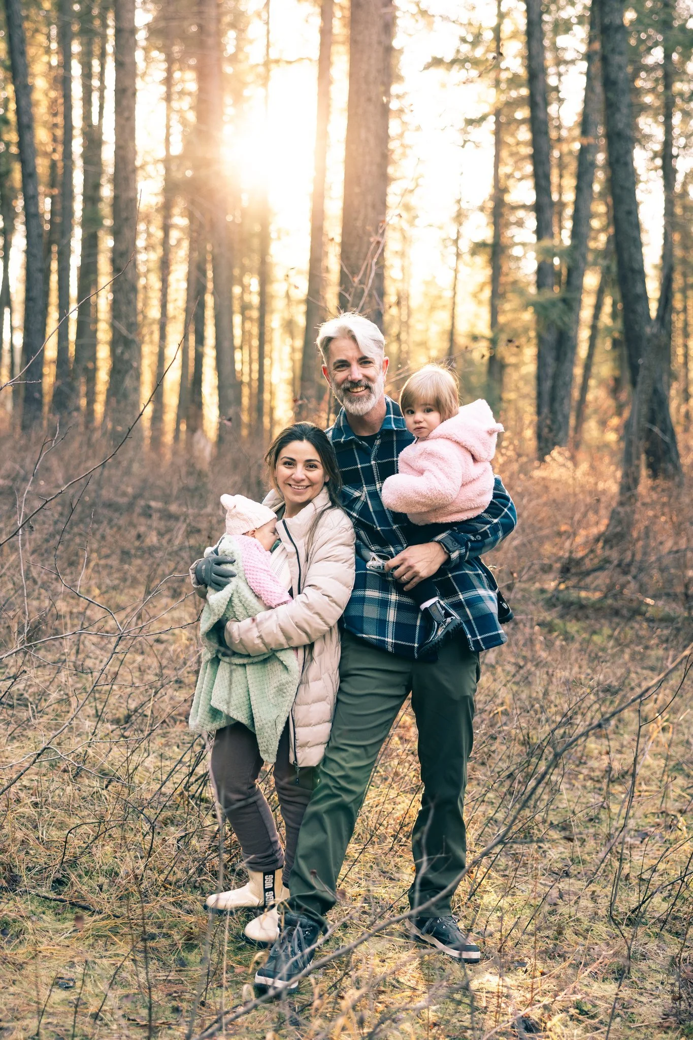 A family of four, including a man, woman, and two young children, in a forest with tall trees and sunlight streaming through the branches.