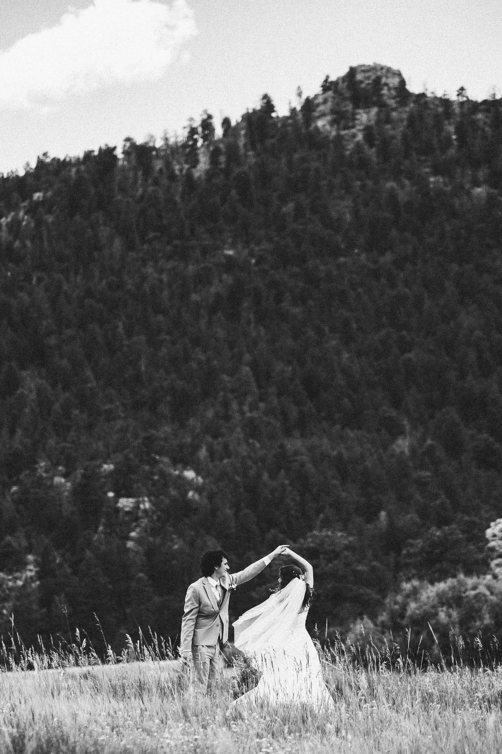 A black and white photo of a wedding couple dancing in a field with tall grass, with trees and a mountain in the background.