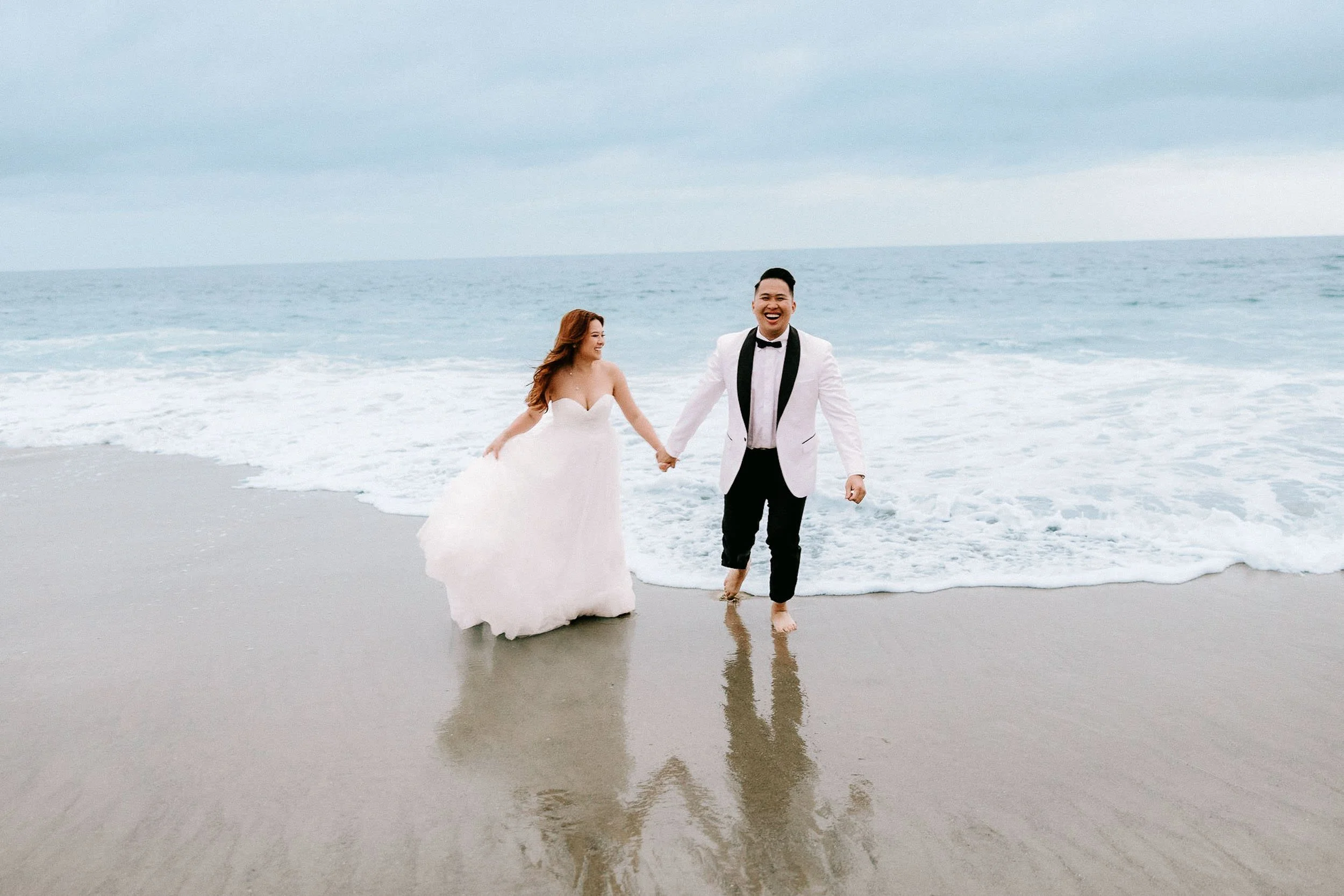 A bride and groom are walking hand in hand along the shoreline of a beach, wearing wedding attire. The bride is in a white wedding dress and the groom is in a tuxedo with a black bow tie. They are smiling and laughing, with the ocean waves reaching their feet under a cloudy sky.